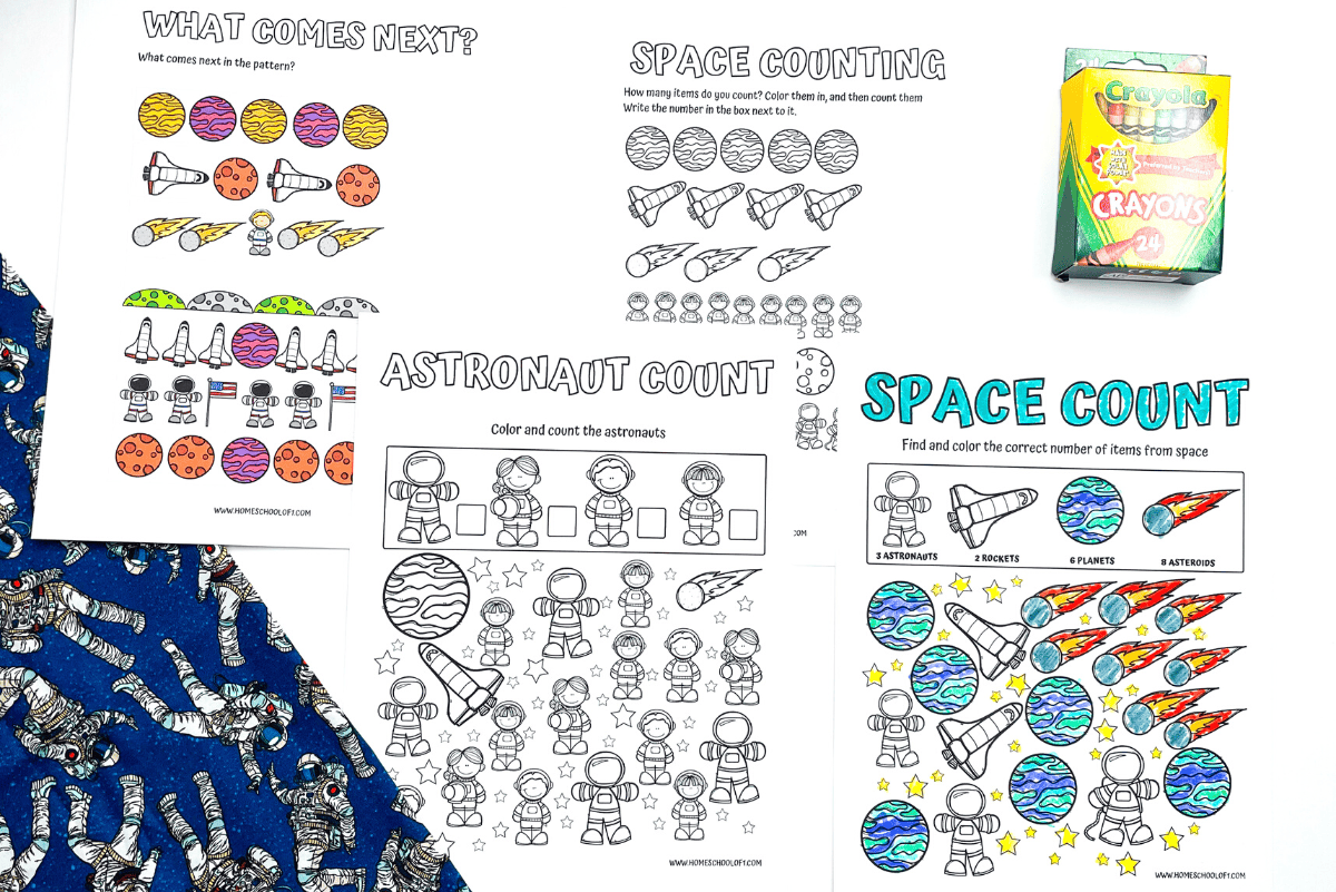 A set of preschool space-themed coloring and counting worksheets featuring rockets, astronauts, planets, and patterns. Some pages are partially colored, and a 24-pack of Crayola crayons sits nearby. A navy blue fabric with astronaut illustrations is partially visible.