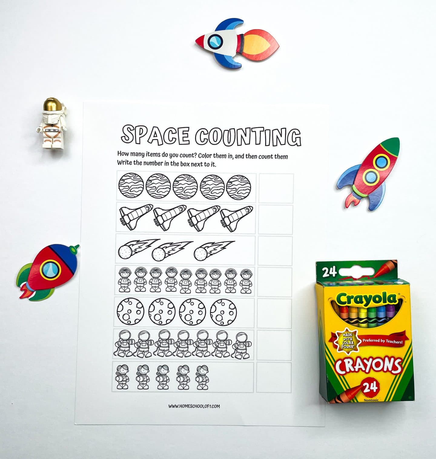 A black-and-white preschool worksheet titled "Space Counting" designed to help children count and color different space-themed items including planets, rockets, meteors, astronauts, and moons. Kids are prompted to count each group and write the total in the boxes provided. Colorful space-themed toys and a 24-pack of Crayola crayons are placed around the worksheet.