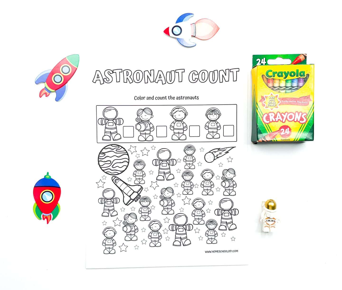 A black and white preschool worksheet titled "Astronaut Count" featuring various cartoon astronauts to color and count, with boxes to tally each type. Surrounding the worksheet are a 24-pack of Crayola crayons, colorful rocket cutouts, and a small astronaut toy.