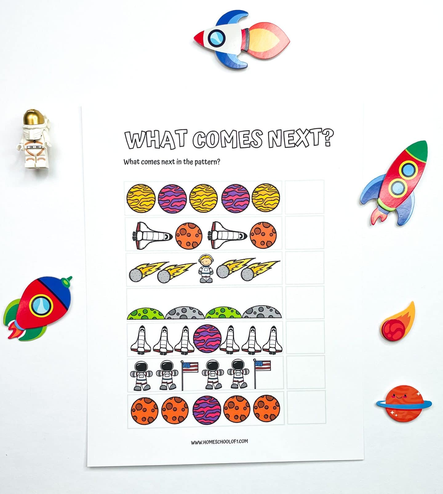A colorful preschool worksheet titled "What Comes Next?" featuring six rows of space-themed pattern sequences with blank spaces at the end for students to predict and complete the patterns. The page is surrounded by small toy rockets, a LEGO astronaut, and space-themed stickers.