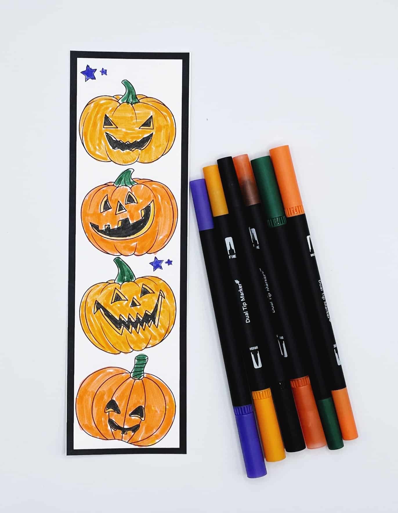 A Halloween-themed bookmark featuring four hand-colored jack-o'-lanterns stacked vertically, with purple stars as accents. Next to the bookmark are five dual tip markers in Halloween colors: purple, yellow, black, green, and orange.