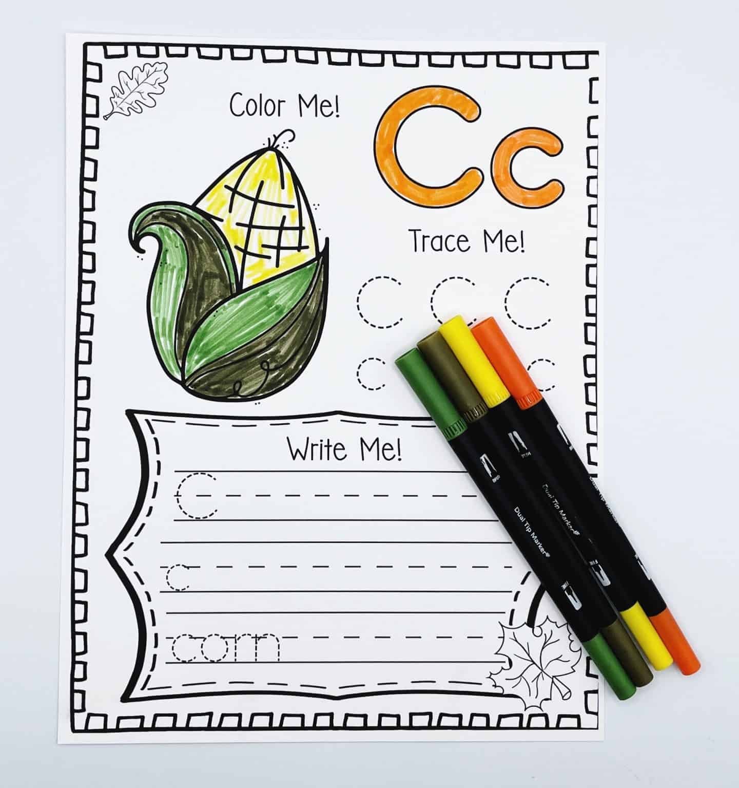 Fall alphabet worksheet for the letter C featuring a corn illustration to color, dotted lines for tracing uppercase and lowercase C, and practice lines to write the word "corn." Four fall-colored markers lie beside the worksheet to complete the autumn theme.







