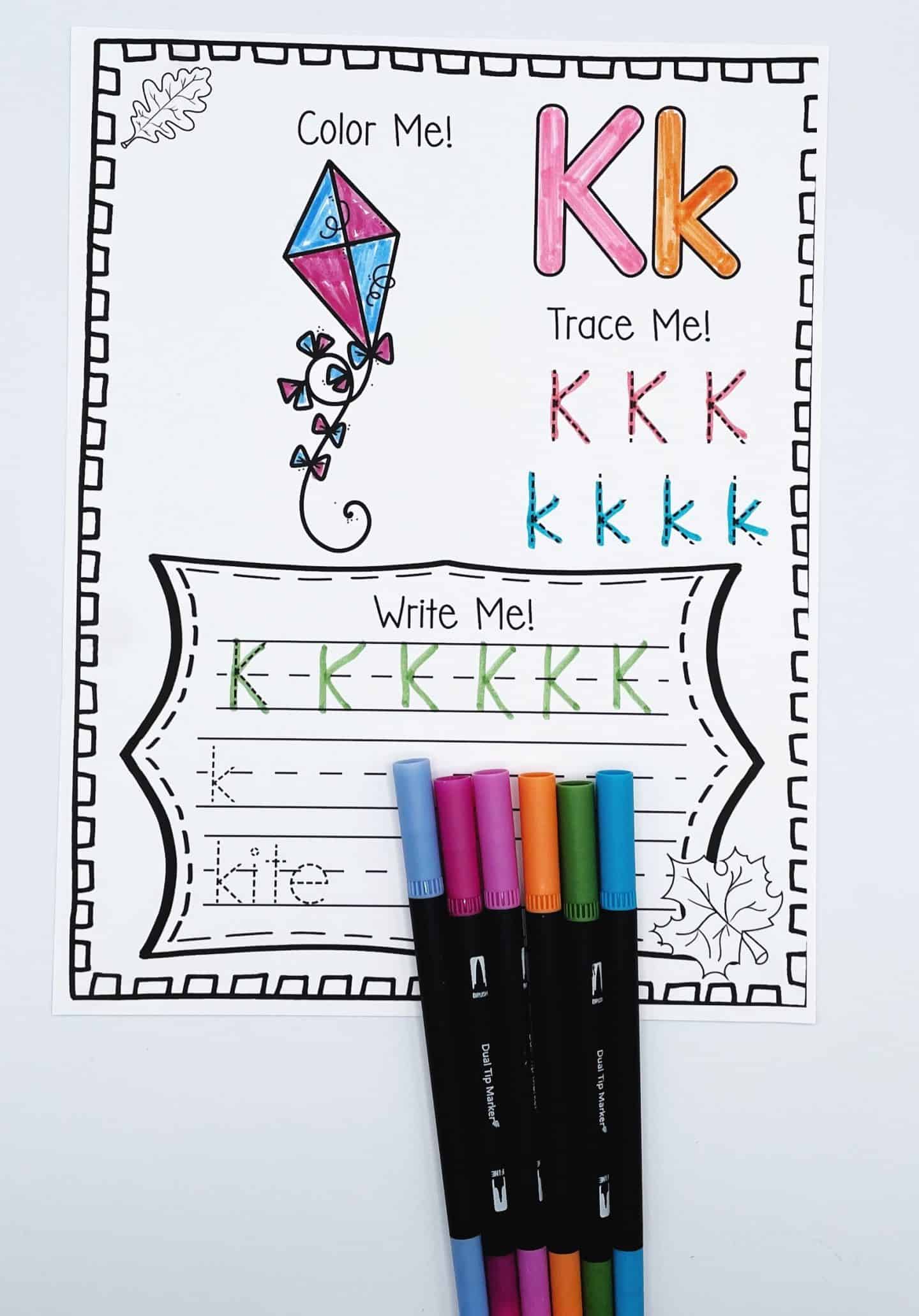 Fall alphabet worksheet for the letter K featuring a kite illustration to color, uppercase and lowercase K tracing practice, and handwriting lines for the word “kite.” A set of colorful markers lies across the bottom, highlighting this engaging early literacy activity.