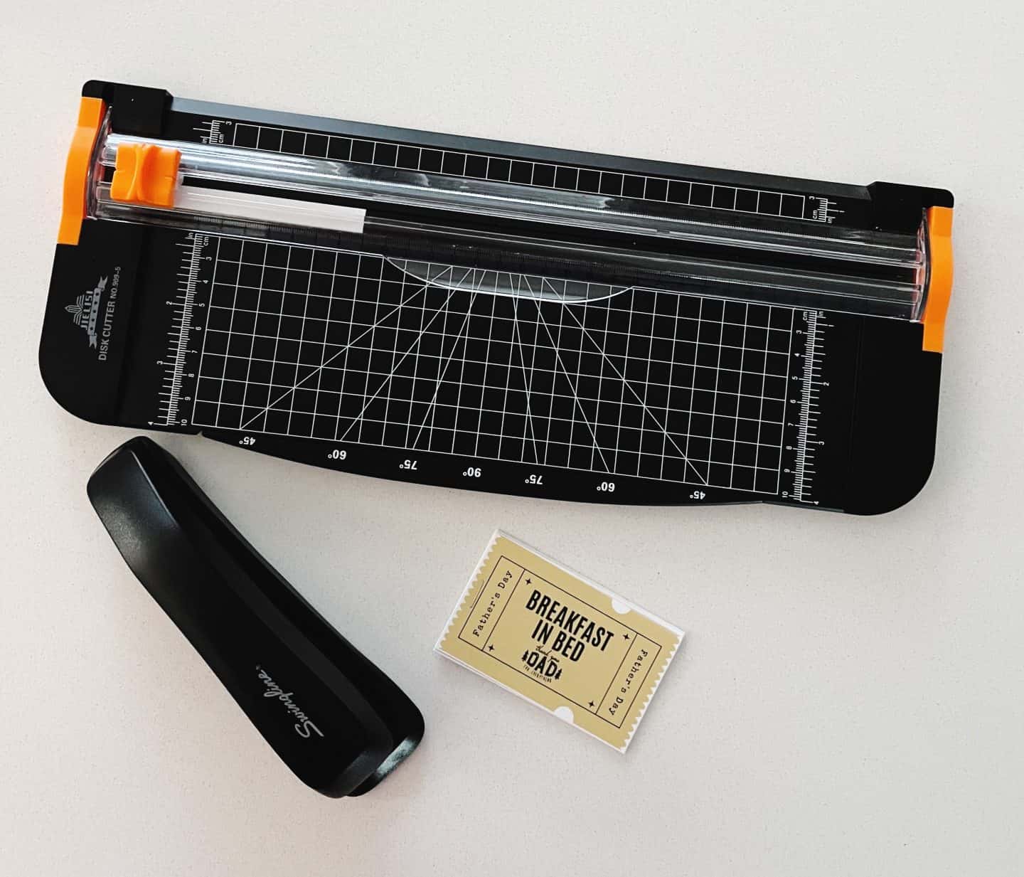 A paper trimmer and black Swingline stapler sit on a white surface beside a freshly cut Father’s Day coupon that says “Breakfast in Bed.” The coupon has a yellow background with black text and a decorative border, suggesting a DIY gift prep in progress.