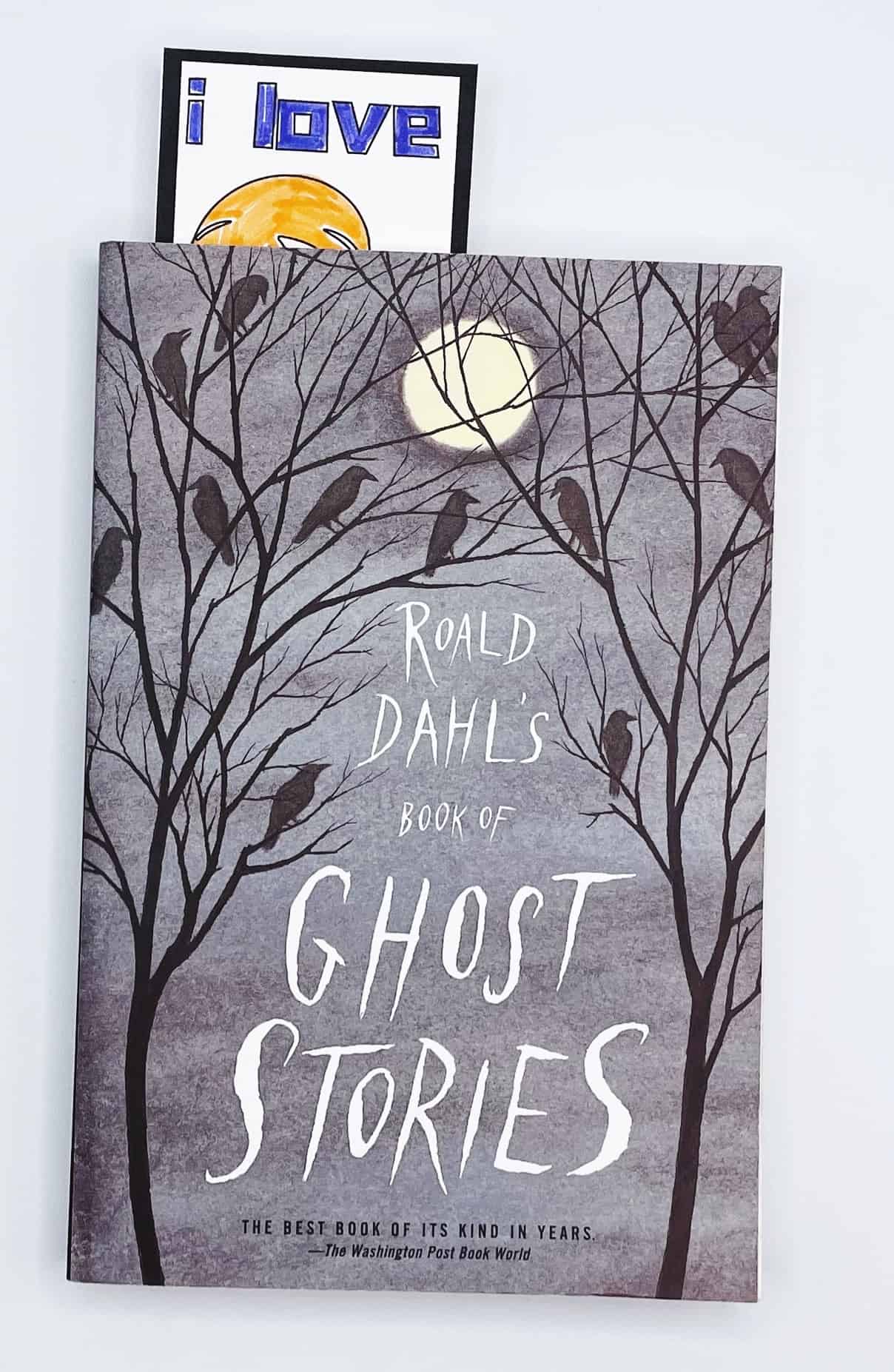 A partially visible Halloween-themed bookmark that says “i love” with a jack-o'-lantern illustration, tucked into the top of a book titled Roald Dahl’s Book of Ghost Stories. The book cover features a spooky night scene with a full moon, bare trees, and perched crows.