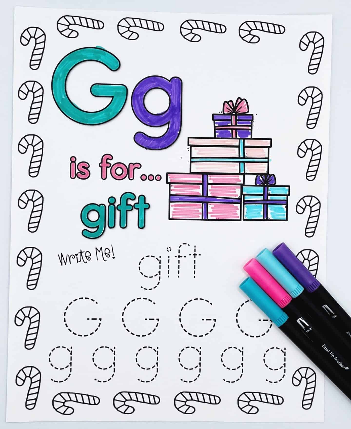 Christmas alphabet worksheet for the letter G, with the phrase “G is for… gift.” The page features a colorful stack of wrapped presents and includes traceable writing practice for the word "gift" and both uppercase and lowercase G’s. The border is decorated with candy canes, and four markers are laid out in the corner.