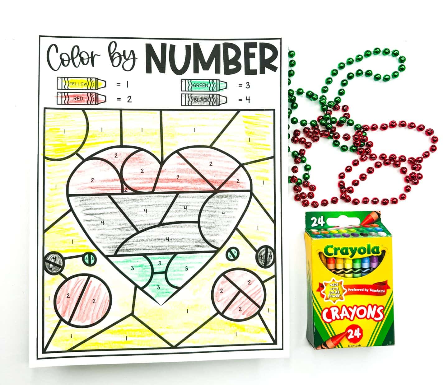 A Juneteenth color-by-number worksheet featuring a large heart and circular shapes, colored using yellow, red, green, and black crayons according to a numbered key at the top. The background is mostly yellow, with the heart filled in red, black, and green segments. The worksheet is placed next to festive red and green beaded necklaces and a 24-pack of Crayola crayons.