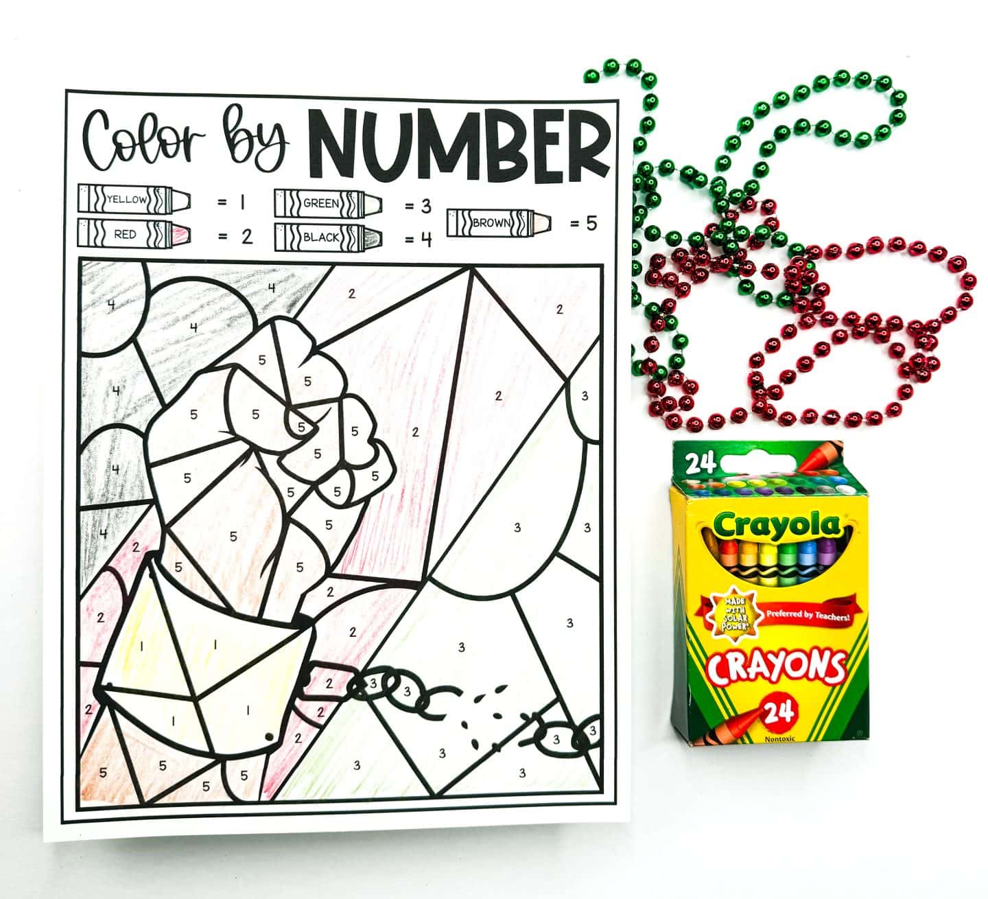 Juneteenth color-by-number worksheet featuring a raised clenched fist breaking free from chains, partially colored using the key at the top which includes yellow, red, green, black, and brown crayons labeled 1 through 5. The fist and surrounding elements are filled in lightly with brown, red, yellow, and green. Red and green bead necklaces and a 24-count box of Crayola crayons are placed beside the worksheet.