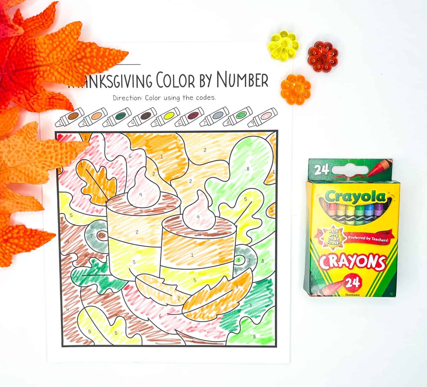 A Thanksgiving color-by-number worksheet featuring two mugs topped with whipped cream, surrounded by autumn leaves in shades of orange, green, and yellow. The image is fully colored using a crayon-coded key above the illustration. A 24-pack of Crayola crayons and festive fall decorations, including faux leaves and plastic flowers, are arranged nearby.