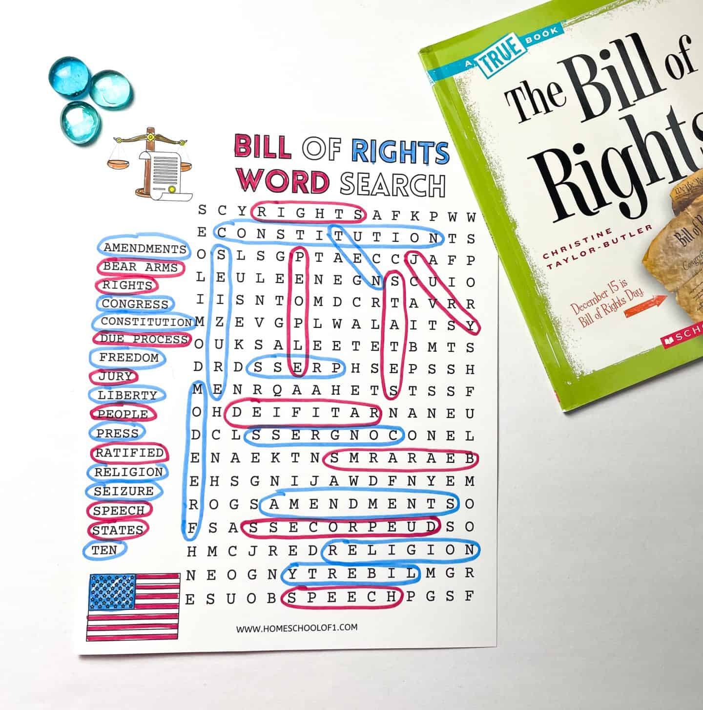 Flat lay of a “Bill of Rights Word Search” worksheet with related terms like “ratified,” “liberty,” and “due process” circled in red and blue; a green children’s book titled The Bill of Rights lies partially on top of the puzzle, and three blue glass gems are placed in the top left corner. The word list and circled answers emphasize vocabulary associated with the U.S. Bill of Rights, with a small American flag graphic at the bottom left.