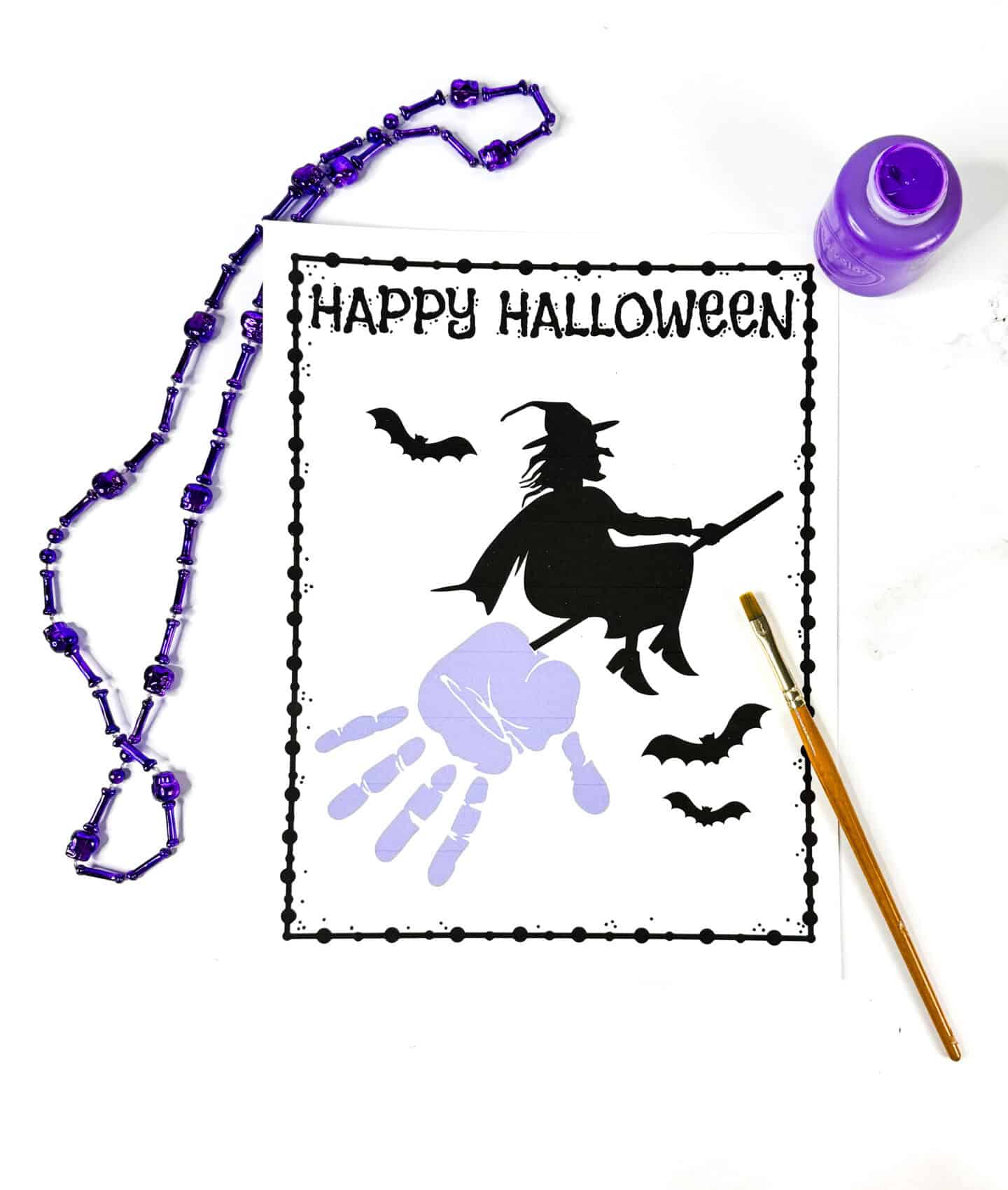 Completed witch handprint craft with purple handprint forming the broom under a flying witch silhouette and Halloween decorations

