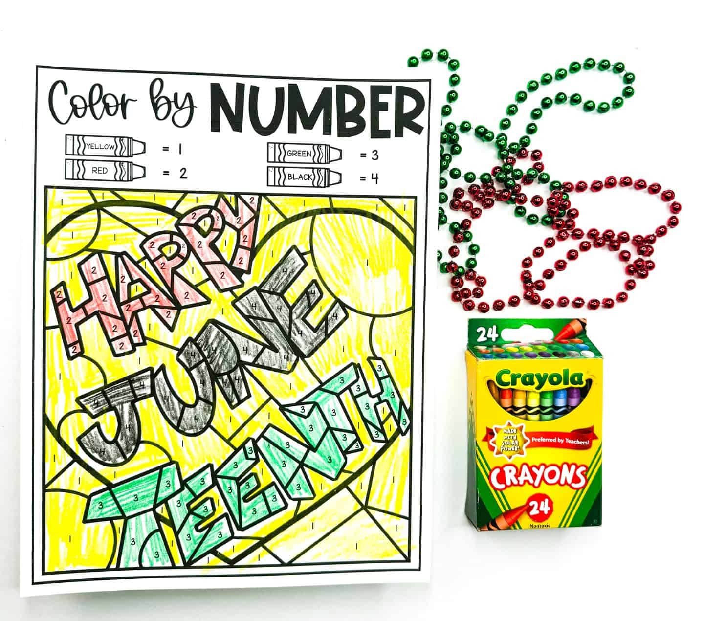 A Juneteenth-themed color-by-number worksheet with the phrase "Happy Juneteenth" filled in using yellow, red, green, and black crayons according to a numbered key. The artwork is placed beside red and green beaded necklaces and a box of 24 Crayola crayons, emphasizing a fun and educational activity for kids to celebrate the holiday.