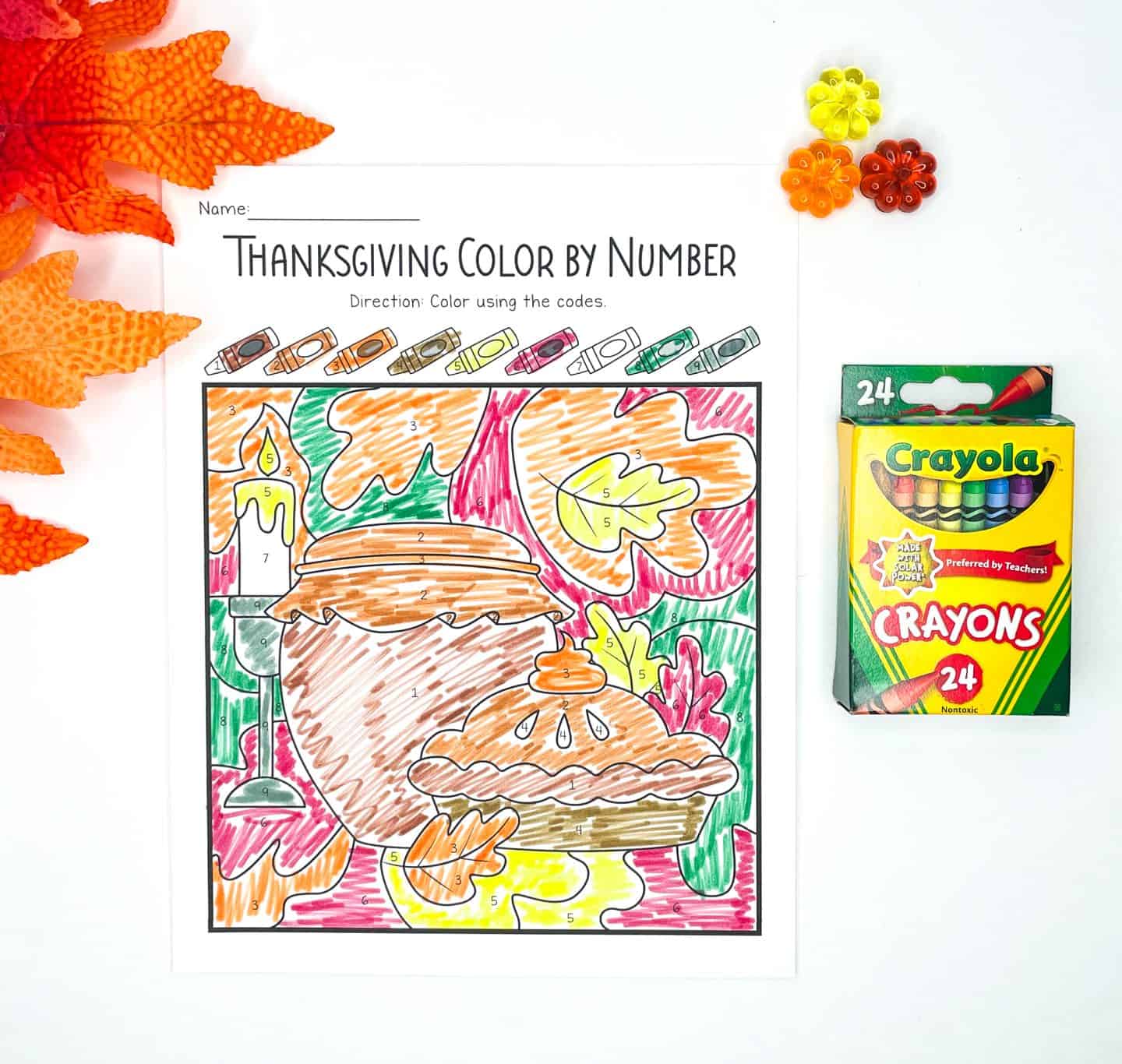 A completed Thanksgiving color-by-number worksheet featuring two pies, a candle, and colorful autumn leaves in the background. The image is filled with bright fall tones using a crayon-coded guide at the top. A box of 24 Crayola crayons and festive fall decorations are arranged alongside the page.

