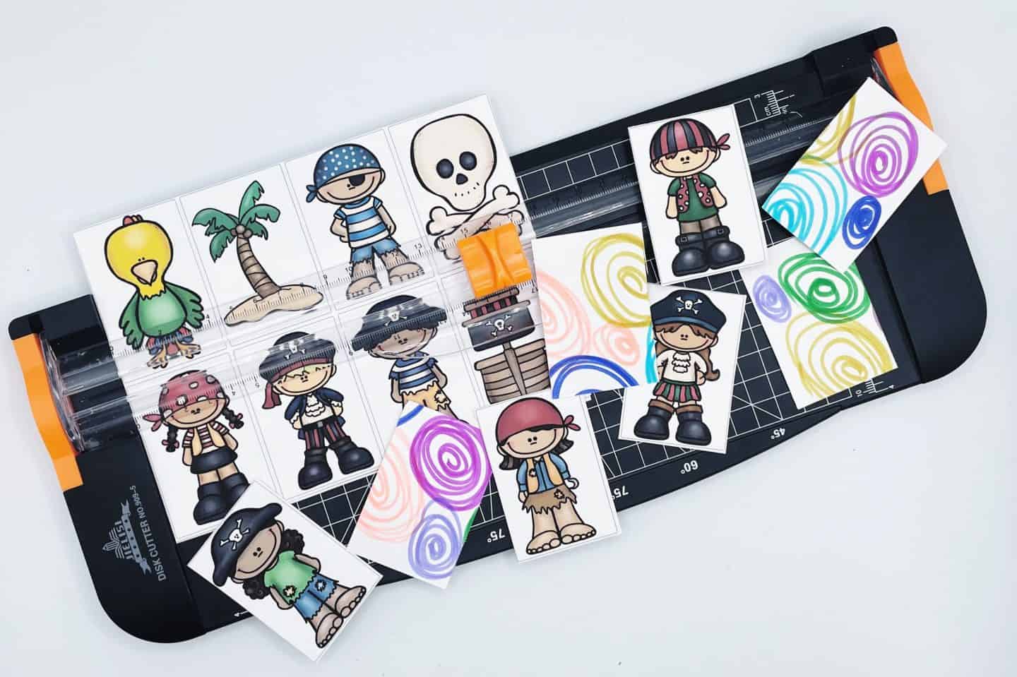 Printable pirate memory cards being cut with a paper trimmer, featuring cartoon images of pirates, a parrot, a palm tree, and a skull. Some cards are flipped to show vibrant swirly backgrounds in blue, green, pink, and yellow, ready for a fun classroom or homeschool matching game.