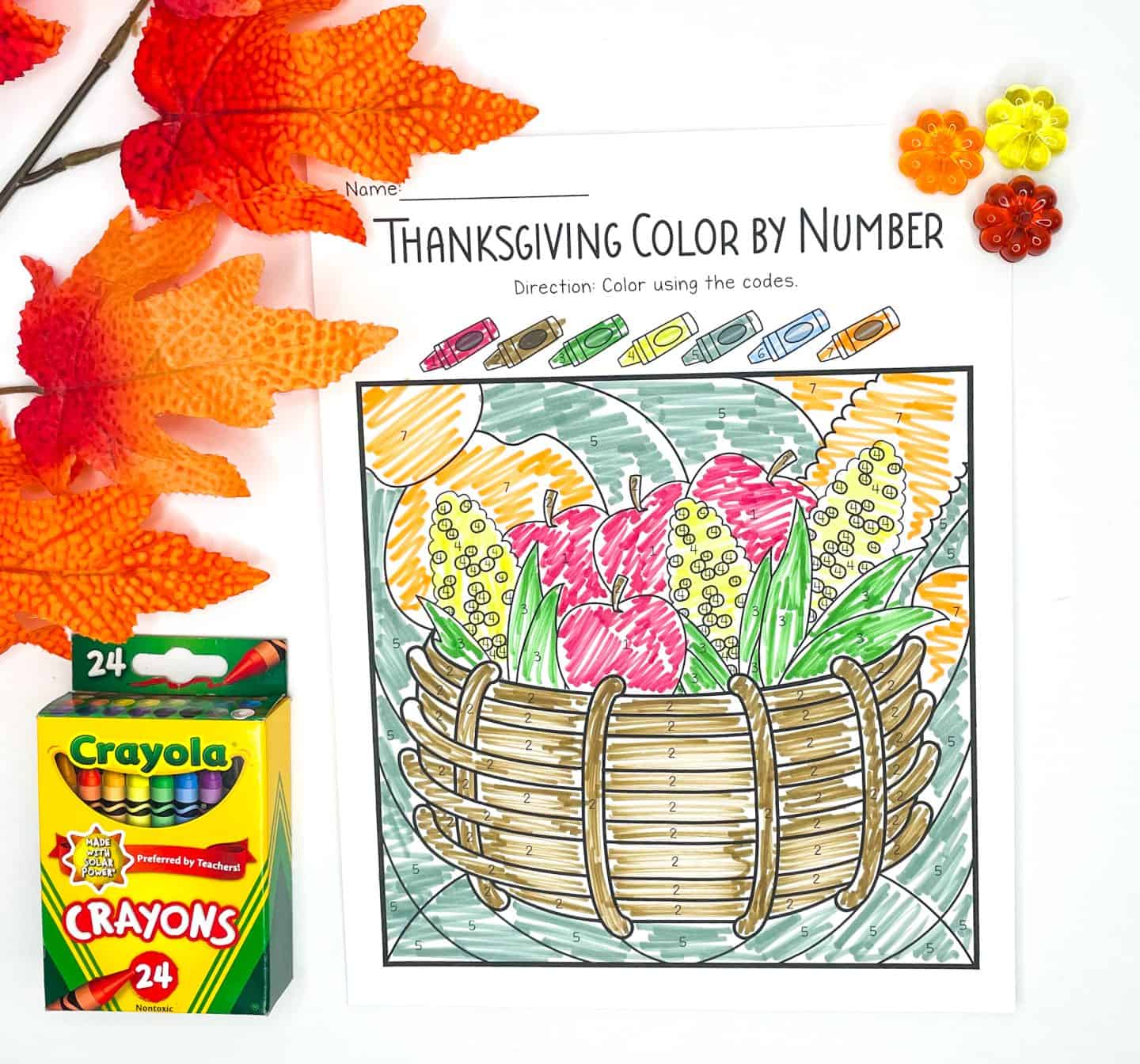 A Thanksgiving color-by-number worksheet is fully colored in, featuring a woven basket filled with apples, corn, pumpkins, and leafy greens. The background and basket are shaded using a numbered color guide at the top, with a box of 24 Crayola crayons and decorative autumn leaves and flowers placed nearby for a festive touch.