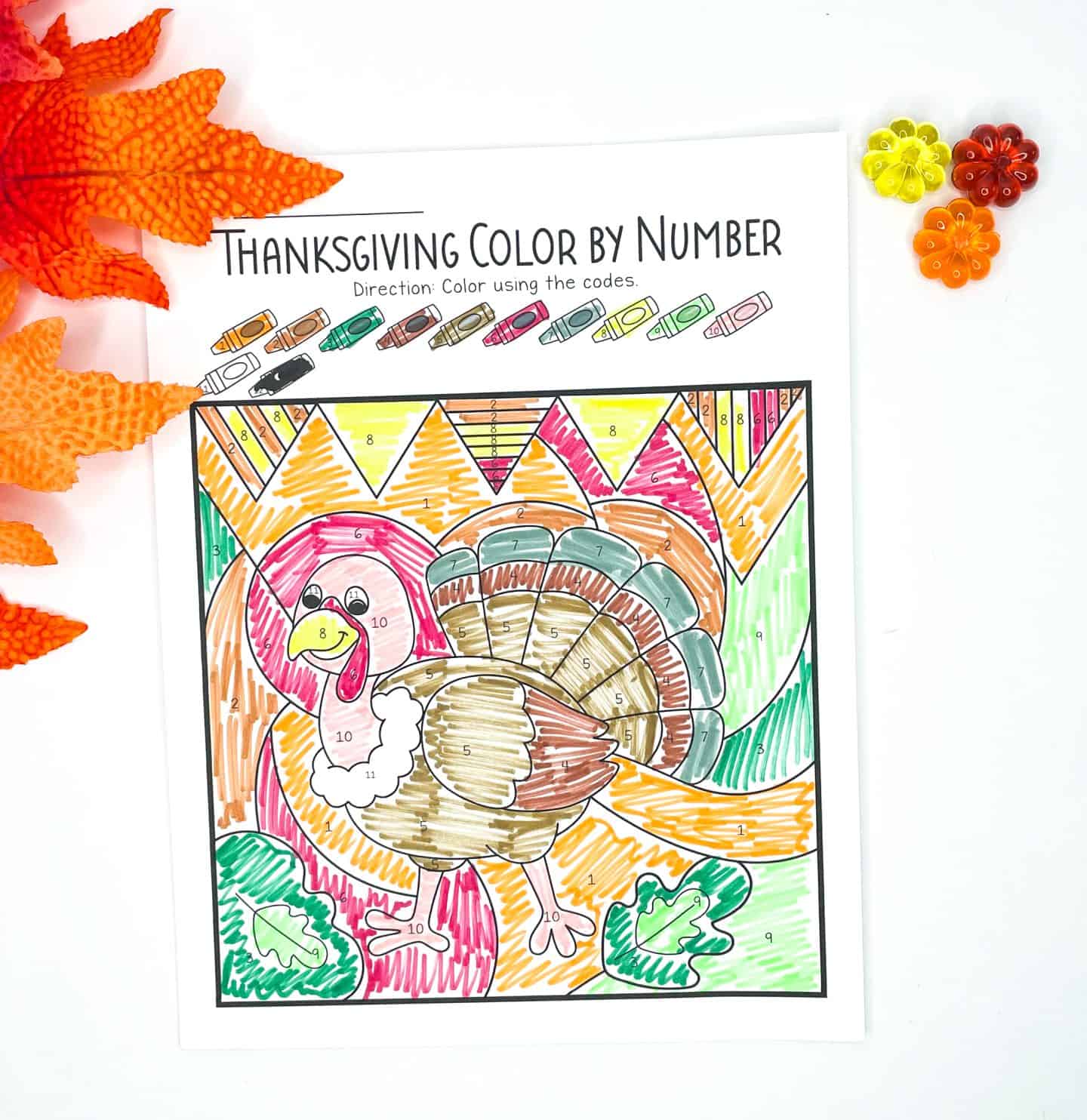 A completed Thanksgiving color-by-number worksheet featuring a brightly colored turkey against a festive background of patterned banners and fall leaves. The image is filled in with colors like red, brown, yellow, green, and orange, following the numbered code at the top. Decorative fall leaves and plastic flowers add a seasonal touch to the scene.