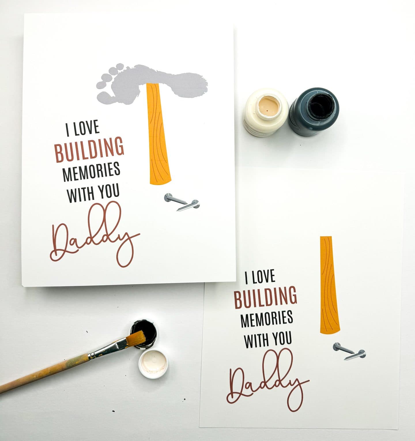 Two DIY Father's Day cards with a construction theme are shown. Each card displays the phrase "I LOVE BUILDING MEMORIES WITH YOU Daddy" with the word "BUILDING" highlighted in brown. The top card has a hammer illustration made from a wooden handle and a gray footprint as the hammerhead, alongside two drawn nails. Paint bottles and a paintbrush with black paint suggest a kid's craft setup.