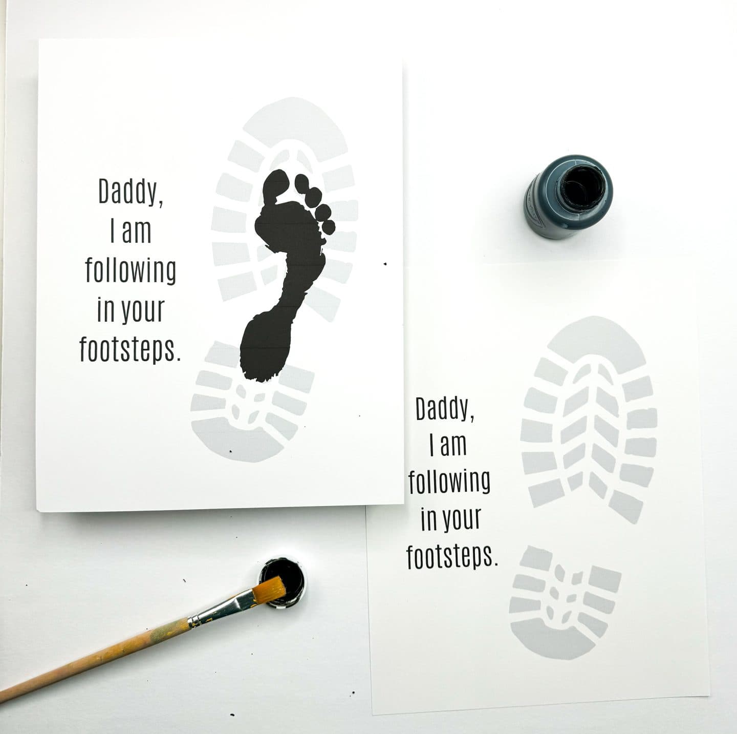 Two Father's Day cards feature a gray boot print and the message, “Daddy, I am following in your footsteps.” One card has a black child’s footprint stamped inside the boot print, representing a symbolic gesture of admiration. Nearby are black paint, a brush, and a paint bottle, suggesting a hands-on craft project for kids.