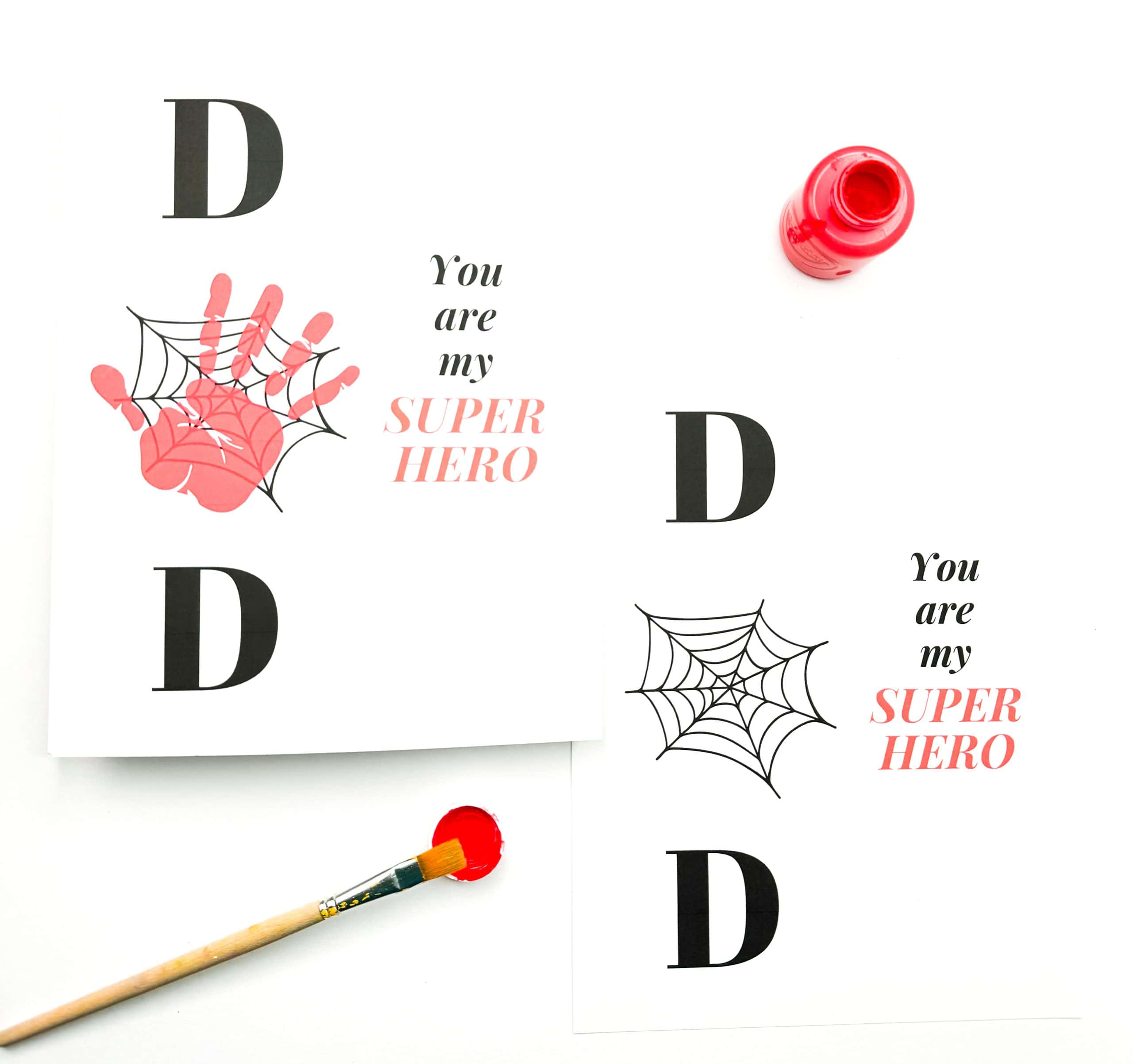 Two DIY superhero-themed Father's Day cards are shown. One card features the word "DAD" with the center "A" replaced by a red handprint overlaid with a black spider web, next to the phrase "You are my SUPER HERO" in black and red font. The second card mirrors the layout but uses only a spider web illustration instead of a handprint. A red paint bottle and brush with red paint sit nearby, hinting at a kid-friendly craft activity.