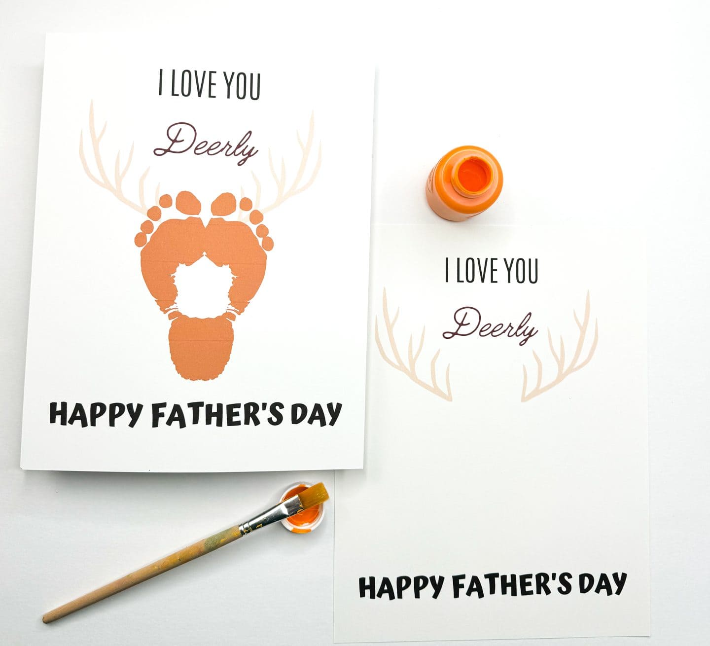 Two Father's Day cards display antlers and the punny message “I LOVE YOU Deerly” above “HAPPY FATHER’S DAY.” One card includes two orange footprints forming the face of a deer between the antlers, while the other is blank and ready for prints. An orange paint bottle and brush are nearby, making this a playful DIY craft for kids.