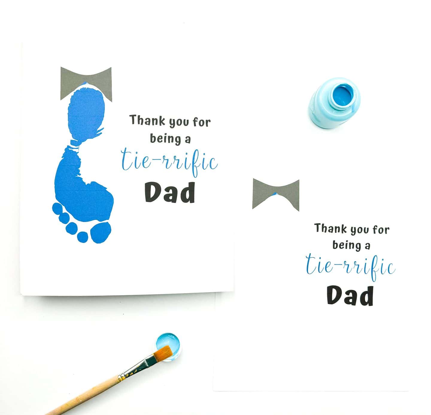 Two handmade Father's Day cards are shown with a bowtie design and the message “Thank you for being a tie-rrific Dad.” One card features a blue footprint below the bowtie, mimicking the shape of a tie, while the other has space left for the footprint. Light blue paint and a brush are nearby, suggesting this is a kid-friendly craft activity.