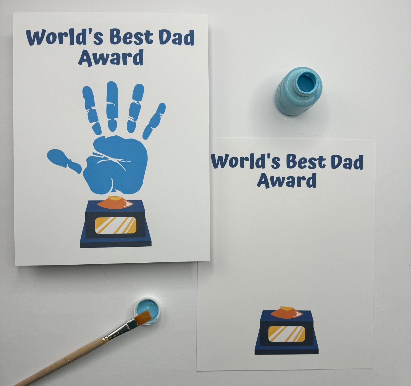 Two Father’s Day craft cards feature the phrase “World’s Best Dad Award” above a trophy illustration. One card includes a blue handprint above the trophy, symbolizing a child's personal touch, while the other is blank and ready for a handprint. A blue paint bottle and brush with light blue paint are placed nearby, highlighting this as a fun DIY gift project for kids.
