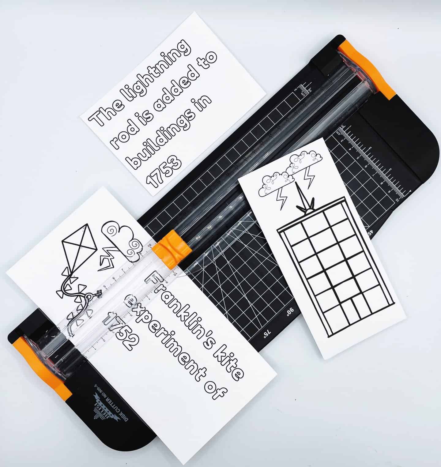 A paper trimmer is shown in use with educational Benjamin Franklin-themed printables, including a card that reads “Franklin’s kite experiment of 1752” alongside a drawing of a kite and lightning, and another card that says “The lightning rod is added to buildings in 1753” with a sketch of a struck building. The setup is part of a hands-on history lesson.