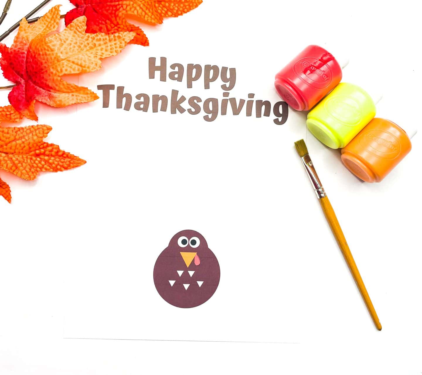 A Thanksgiving craft template displays the phrase "Happy Thanksgiving" in brown text at the top. Below it is a simple cartoon turkey with a round dark brown body, white triangular feathers on its belly, wide eyes, a yellow beak, and a red wattle. To the right are red, yellow, and orange paint pots with a paintbrush, and on the left, artificial orange fall leaves add a festive touch.