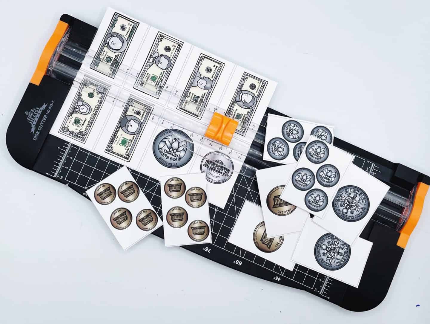 A paper trimmer is being used to cut out pieces from a printable Money Memory Game. The printed pieces include images of U.S. currency such as $1 and $10 bills and various coins like pennies and quarters. Several small rectangular cards with coin illustrations are scattered around the trimmer, ready to be used in the memory game.