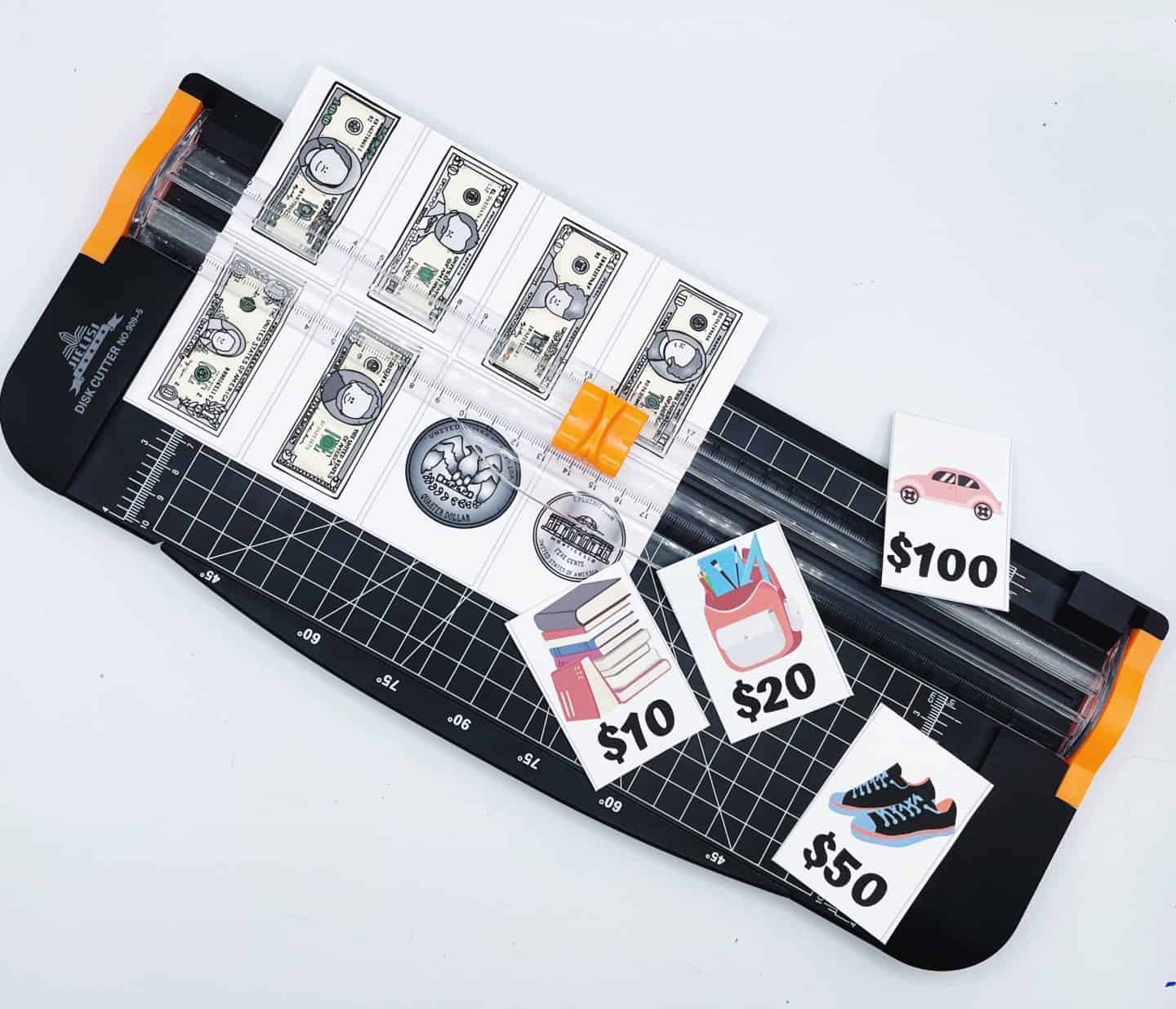 A paper trimmer is shown cutting out money memory game cards featuring illustrated U.S. bills and coins. Beside the trimmer are cut-out cards with images of items like a car labeled $100, sneakers labeled $50, a backpack labeled $20, and books labeled $10, part of an educational game for kids to match money to prices.