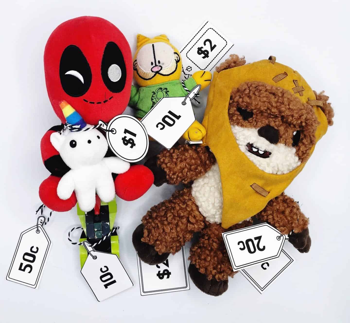 A group of plush toys with attached price tags used for a kids' money learning activity. The toys include a red Deadpool-like character holding a white unicorn plush ($1), a yellow Ewok plush ($2), a green Garfield-style cat (10¢), and a few smaller toys labeled with 10¢, 20¢, and 50¢. The setup teaches basic coin recognition and pricing.


