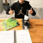 A middle-school student sitting at a table with the Real Science Odyssey Biology 2 textbook, a microscope, and open workbook pages, giving two thumbs up during a homeschool biology lesson.