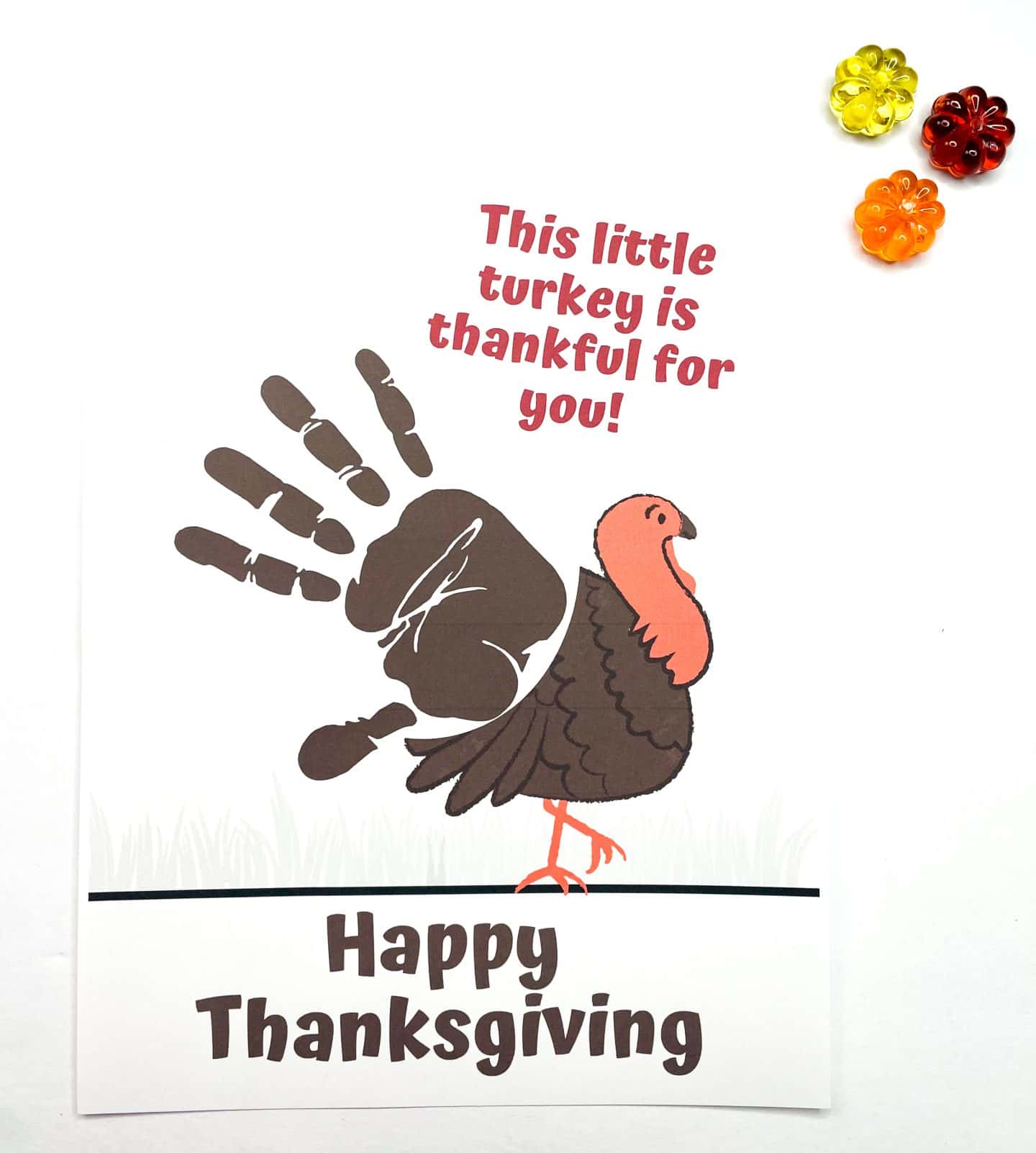 A Thanksgiving-themed craft features the phrase "This little turkey is thankful for you!" in red text above a handprint turkey made with brown paint. The handprint forms the body of the turkey, which is illustrated with a cartoon head and feet. The message "Happy Thanksgiving" is printed at the bottom in bold brown letters. Three small fall-themed glass embellishments—yellow, orange, and red—are placed in the top right corner.
