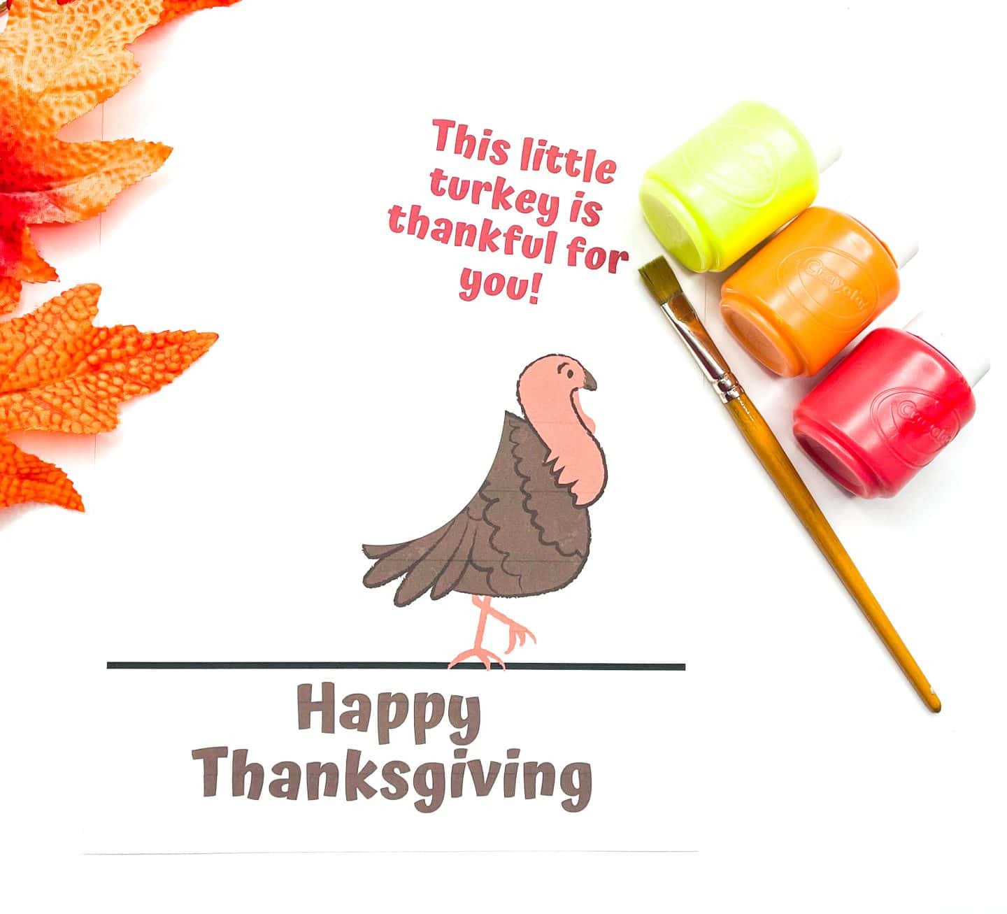 A Thanksgiving craft page featuring a cartoon-style brown turkey standing above the text "Happy Thanksgiving." Above the turkey, red text reads "This little turkey is thankful for you!" The setup includes a paintbrush and small paint containers in yellow, orange, and red, with orange artificial leaves arranged on the left side, suggesting a fun and festive painting activity for kids.