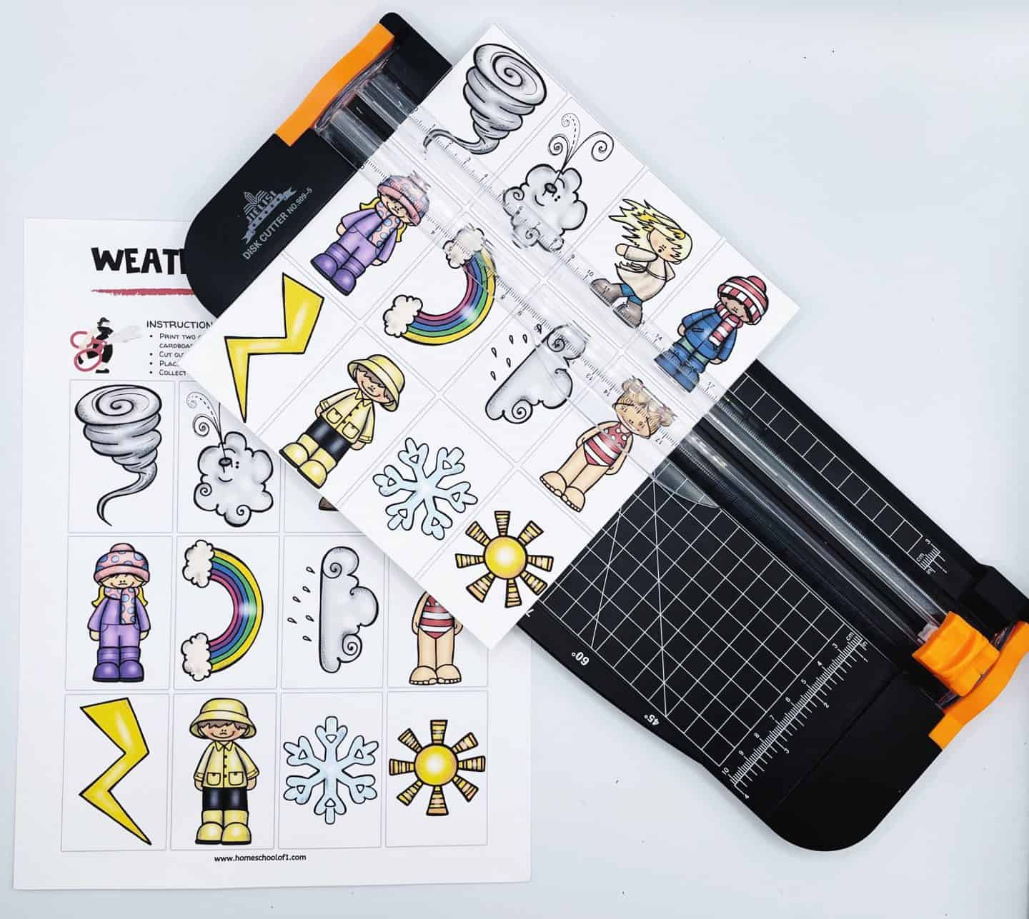 A paper sheet with weather-themed illustrations including a tornado, thunderbolt, rainbow, snowflakes, and sun, is placed on a black paper cutter. The cutter is positioned diagonally over a weather activity worksheet.
