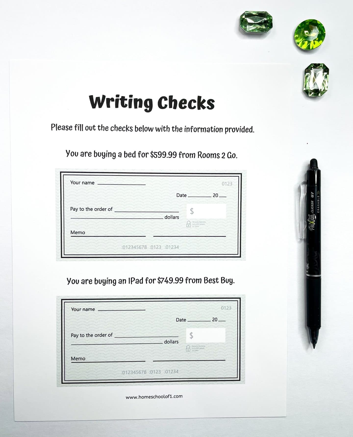 A printable worksheet titled "Writing Checks" includes two practice check templates. The top section prompts the user to fill out a check for $599.99 to Rooms 2 Go for a bed. The bottom section asks for a check to be written for $749.99 to Best Buy for an iPad. Each check includes blank fields for name, date, payee, dollar amount, memo, and signature. Green gem decorations and a black pen are placed beside the sheet.