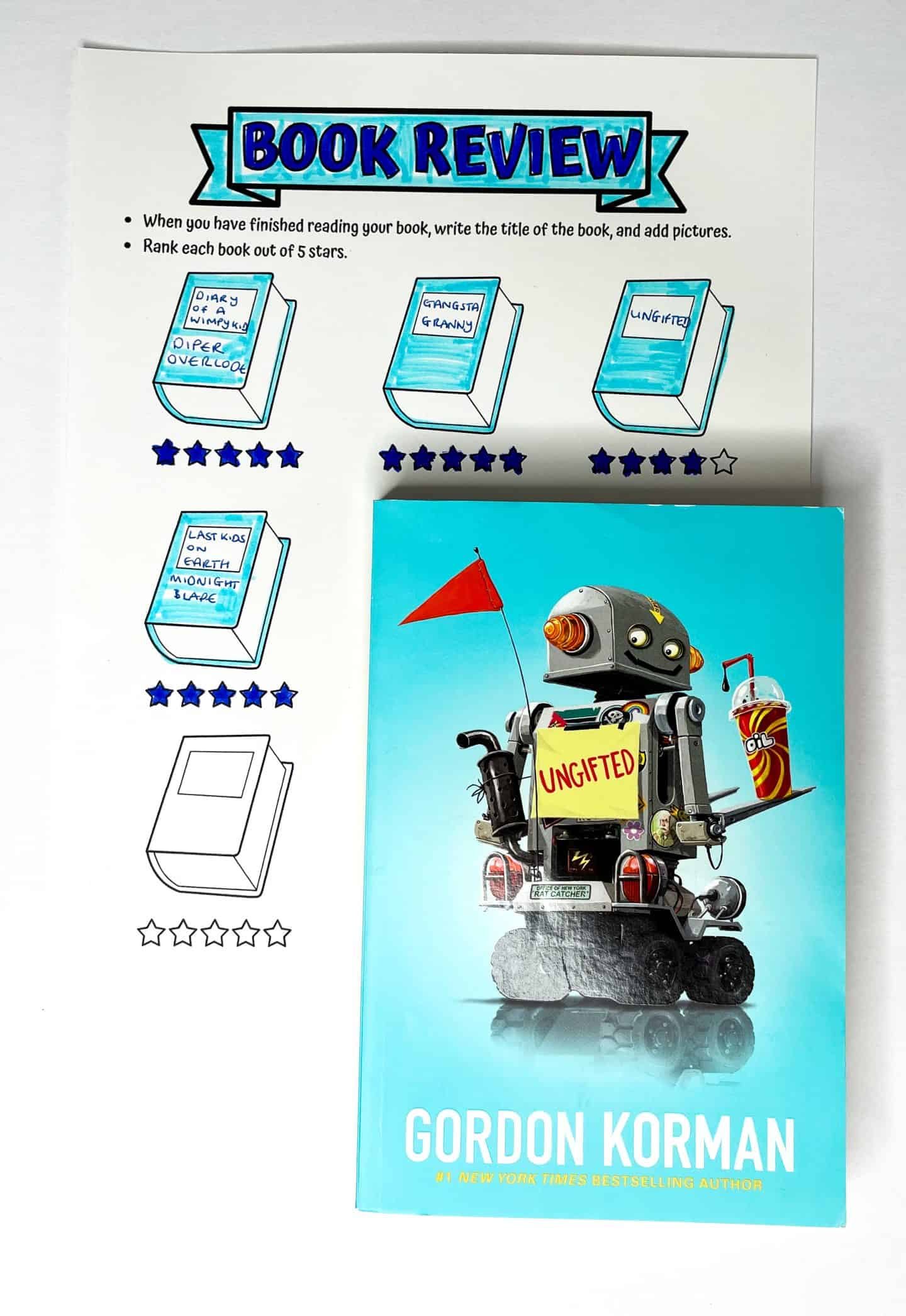 A “Book Review” printable worksheet with five illustrated books, each labeled with a title and star rating. Titles include Diary of a Wimpy Kid: Diper Overlode, Gangsta Granny, Ungifted, and The Last Kids on Earth: Midnight Blade. Four books are rated with five stars, while Ungifted has four stars. Below the worksheet is a physical copy of the book Ungifted by Gordon Korman, featuring a robot on the cover holding a sign that says "Ungifted" and a slush drink.