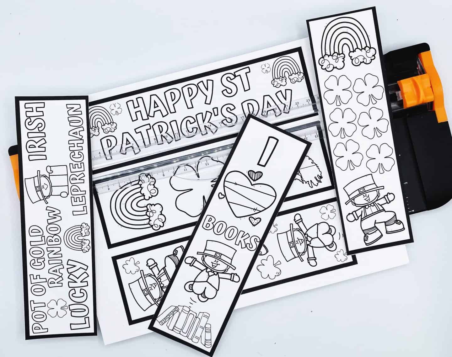 A collection of printable black-and-white St. Patrick’s Day bookmarks laid out beside a paper cutter. Designs include rainbows, shamrocks, leprechauns, the phrase “Happy St. Patrick’s Day,” and “I ? Books.” One bookmark also features themed words like “Pot of Gold,” “Rainbow,” “Lucky,” and “Leprechaun.” These bookmarks are ready to be colored and cut out for festive reading fun.