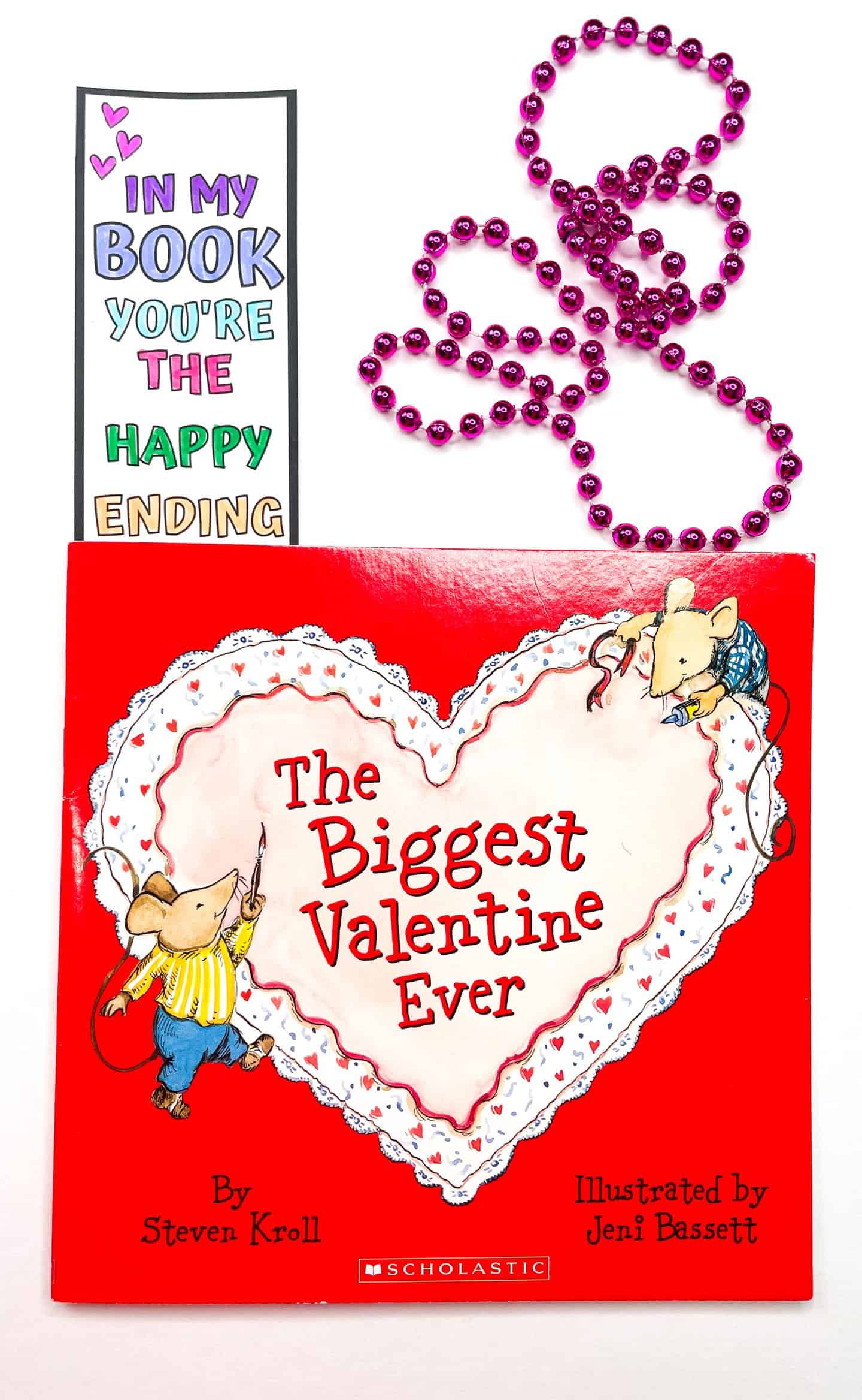 A Valentine’s Day bookmark tucked into the top of the children’s book The Biggest Valentine Ever by Steven Kroll. The bookmark reads “In my book you’re the happy ending” in colorful, hand-colored letters, with purple hearts and a cartoon cupid. A pink beaded necklace is curled beside the book on a white background.