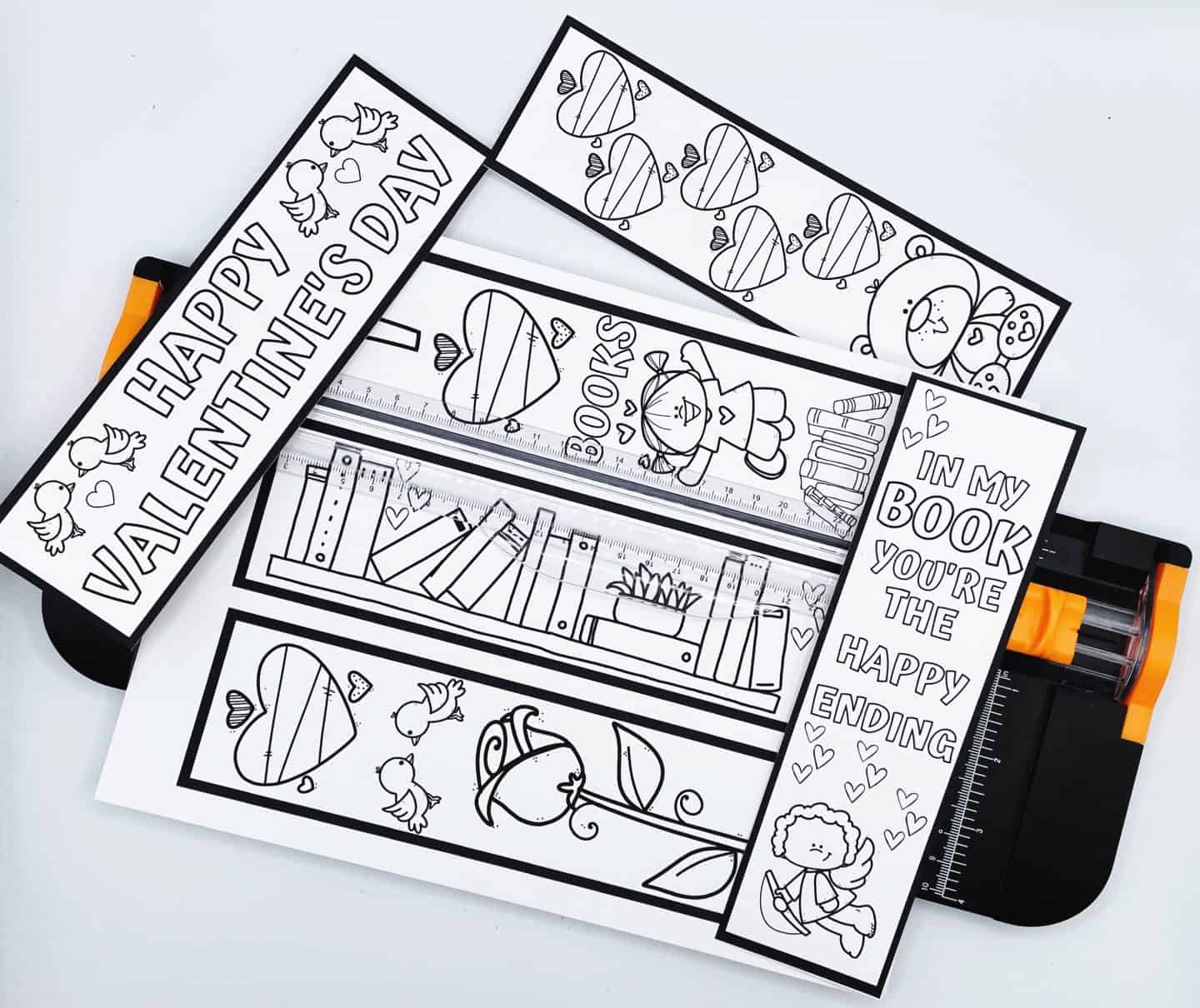 Printable Valentine's Day bookmarks in black and white laid out for cutting, featuring heart designs, lovebirds, roses, books, and sweet phrases like “In my book you’re the happy ending” and “Happy Valentine’s Day.” A paper trimmer is positioned underneath, ready to slice the bookmarks apart. Perfect for coloring and gifting.