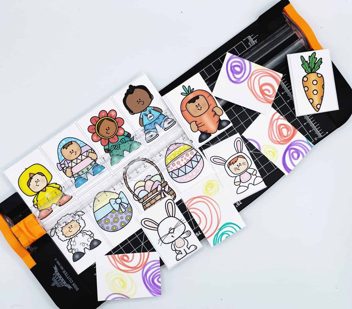Close-up of an Easter memory game in progress, with character cards being cut using a paper trimmer. The cards feature colorful cartoon illustrations of kids dressed as bunnies, flowers, carrots, and more, along with Easter baskets, eggs, and lambs. The backs of the cards display vibrant swirls in purple, red, green, and yellow, making it a cheerful and hands-on craft