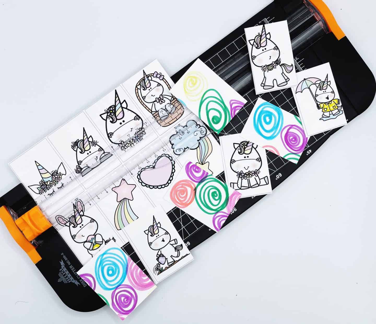 A paper trimmer in use with unicorn-themed memory game cards being cut out. The cards feature cute black-and-white unicorn illustrations and colorful swirl-patterned backs. Some cards are fully cut and scattered around the trimmer, ready for gameplay or laminating.