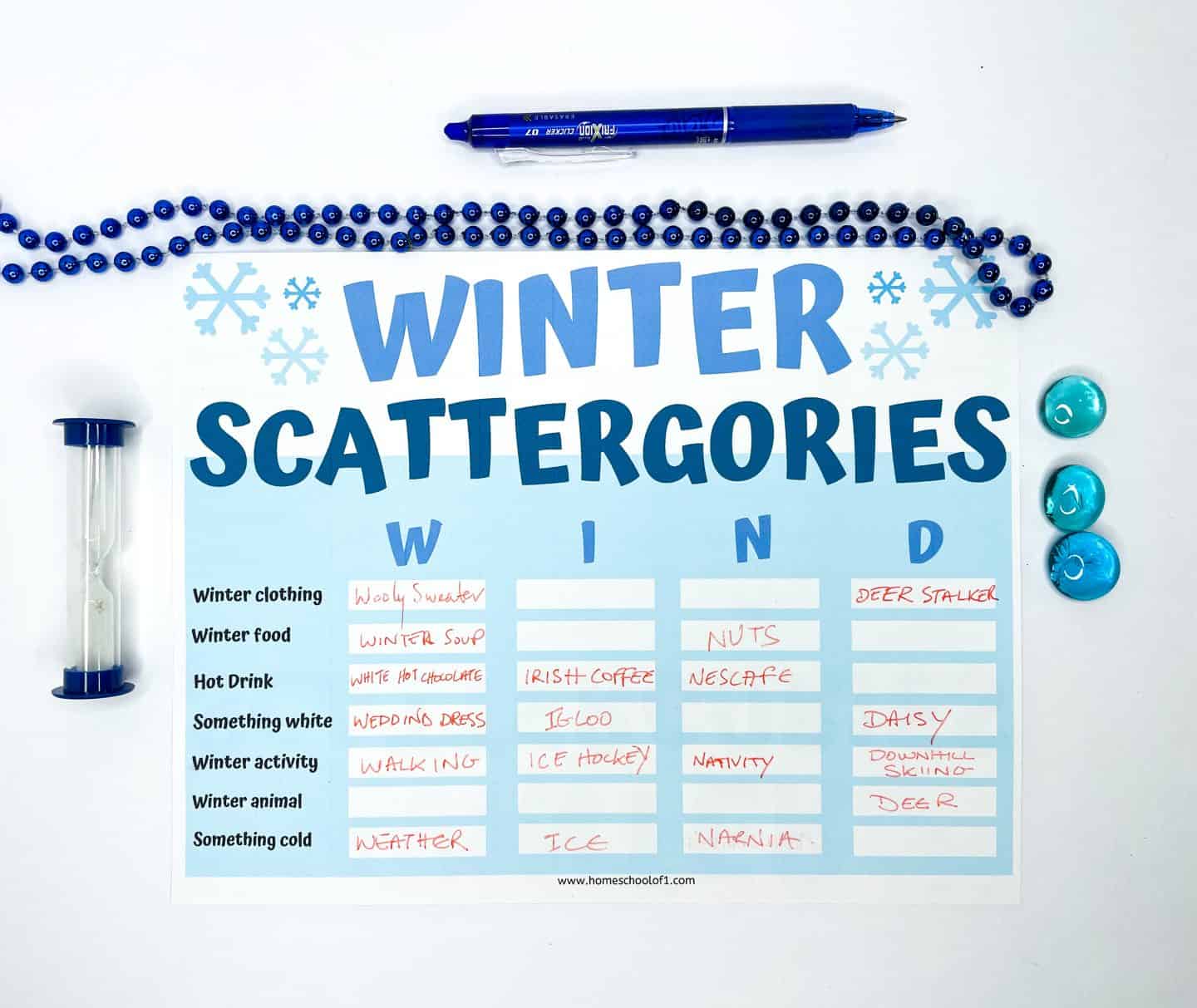A filled-in “Winter Scattergories” game sheet with answers for the letters W, I, N, and D under categories like Winter Clothing, Winter Food, Hot Drink, and Winter Activity. Entries include “Woolly Sweater,” “Irish Coffee,” “Nuts,” “Deer Stalker,” and “Narnia.” The sheet is surrounded by a blue beaded necklace, a blue pen, blue glass game pieces, and a sand timer, all set on a white background.