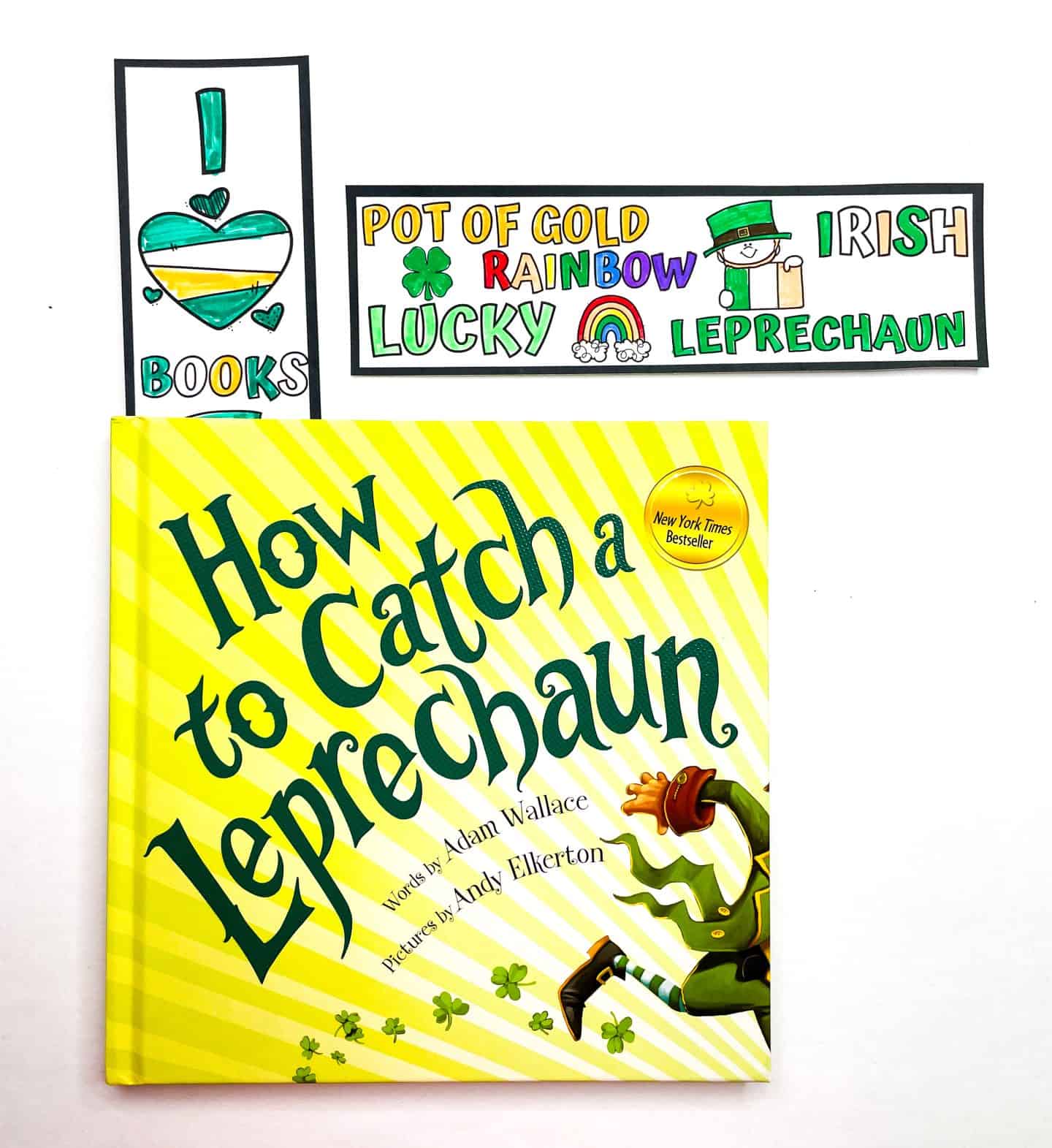 Two colorful St. Patrick’s Day bookmarks sit above the bright yellow children’s book How to Catch a Leprechaun by Adam Wallace. One bookmark reads “I ? Books” with a heart colored like the Irish flag, and the other features words like “Pot of Gold,” “Rainbow,” “Lucky,” and “Leprechaun,” with festive drawings of a shamrock, rainbow, and leprechaun.