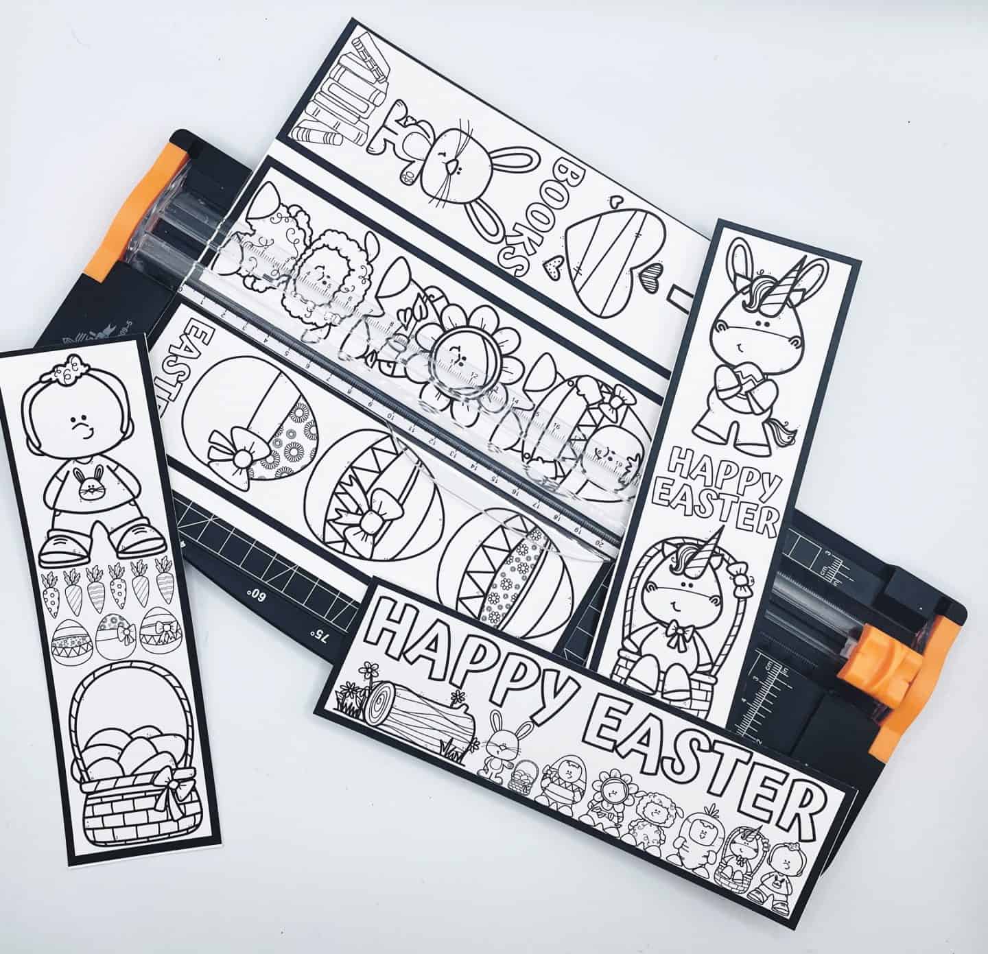Several uncolored Easter bookmarks are being trimmed on a paper cutter. Designs include Easter eggs, bunnies, spring animals, and the phrase “Happy Easter” in bold bubble letters.

