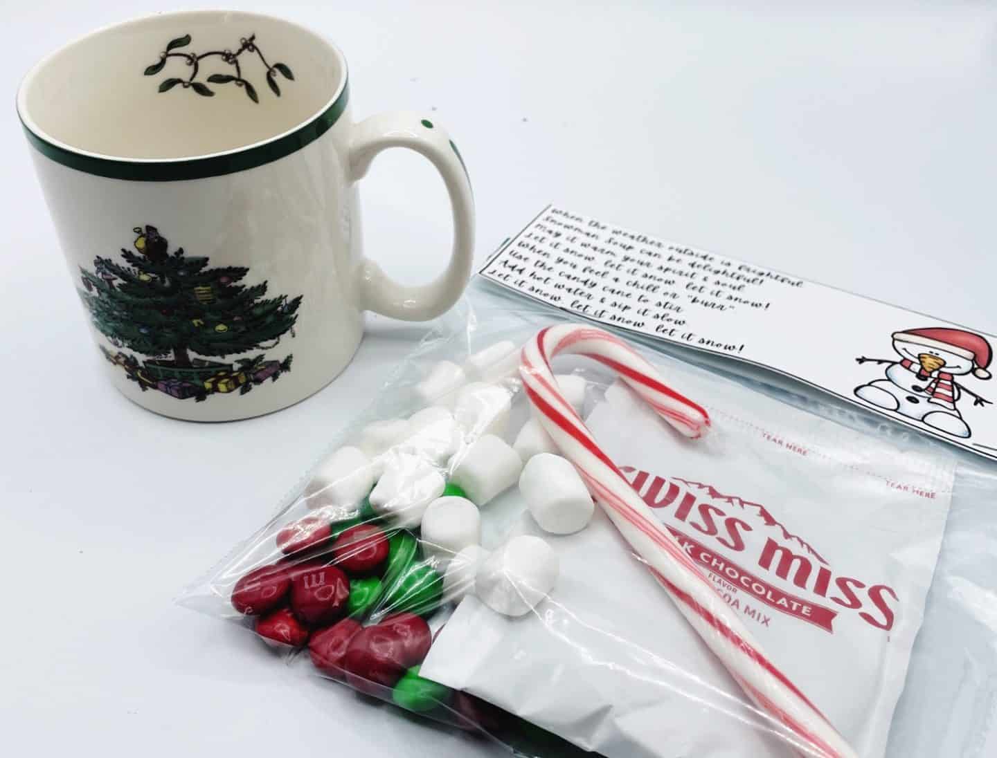 A festive Snowman Soup free printable gift set displayed with a holiday-themed mug featuring a decorated Christmas tree. The treat bag includes a packet of Swiss Miss hot chocolate, mini marshmallows, red and green M&Ms, and a candy cane, topped with a printable poem tag featuring a smiling snowman. Perfect for cozy winter gifts, class treats, or holiday party favors.