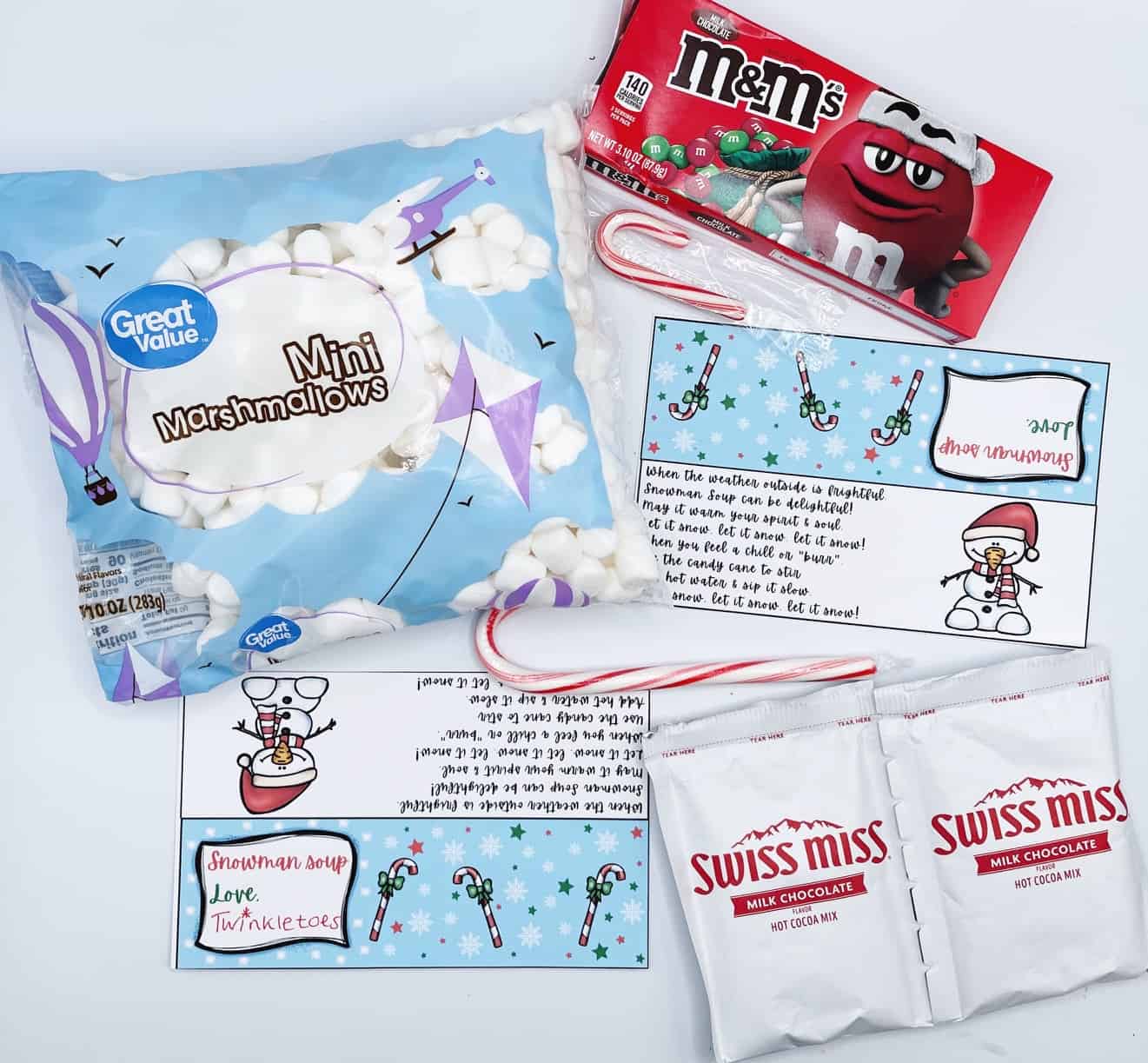  A fun Snowman Soup free printable kit laid out with all the ingredients: a bag of Great Value mini marshmallows, a box of holiday M&M’s, Swiss Miss hot cocoa mix, candy canes, and colorful printable toppers featuring a festive snowman and rhyming instructions. The printables read “Snowman Soup Love, Twinkietoos” and are designed to fold over treat bags for an easy DIY winter gift.