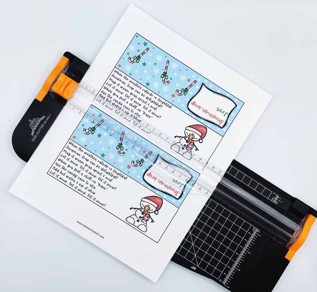 A sheet of Snowman Soup free printable gift tags being trimmed with a paper cutter. Each tag features a festive poem and a cute snowman illustration, along with a winter background of snowflakes and candy canes. Perfect for creating DIY hot cocoa treat bags for Christmas gifts, classroom favors, or holiday parties.