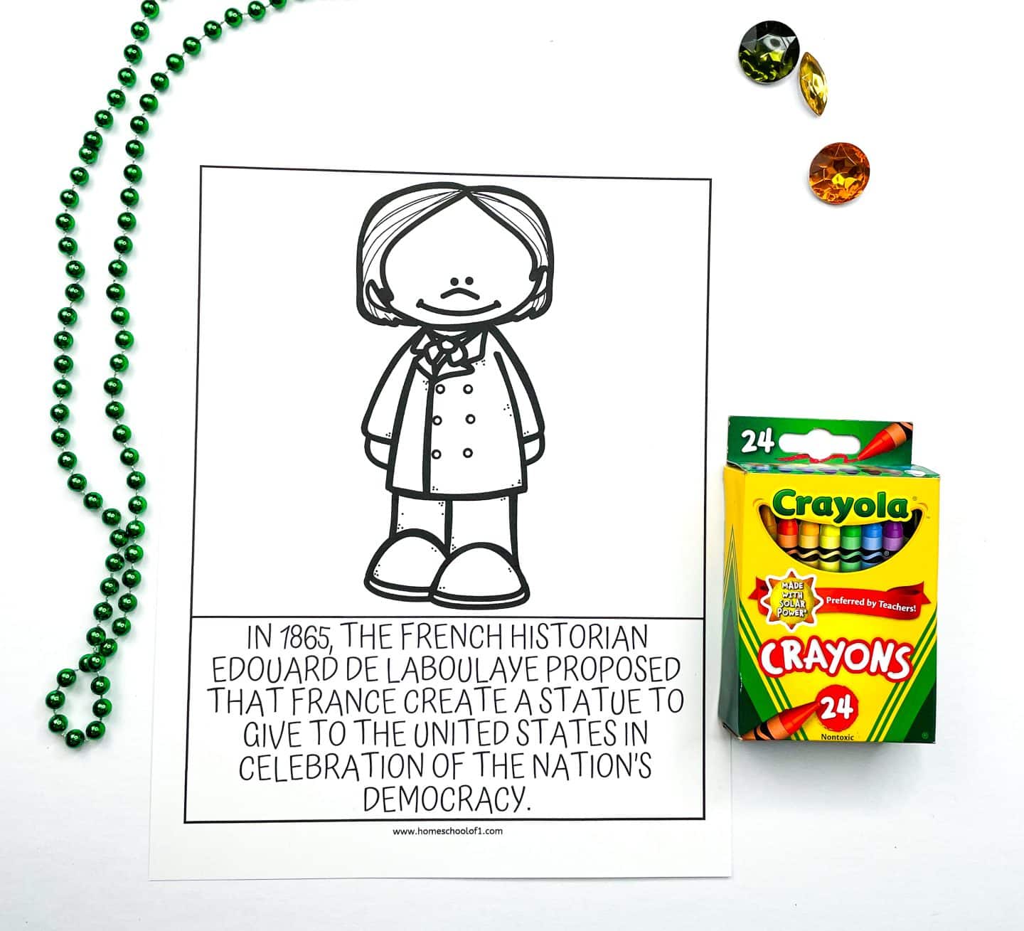 A black and white educational coloring worksheet featuring a cartoon illustration of French historian Edouard de Laboulaye. Below the image, a caption reads: “In 1865, the French historian Edouard de Laboulaye proposed that France create a statue to give to the United States in celebration of the nation’s democracy.” The worksheet is surrounded by a green beaded necklace, three colorful plastic gems, and a box of Crayola crayons.