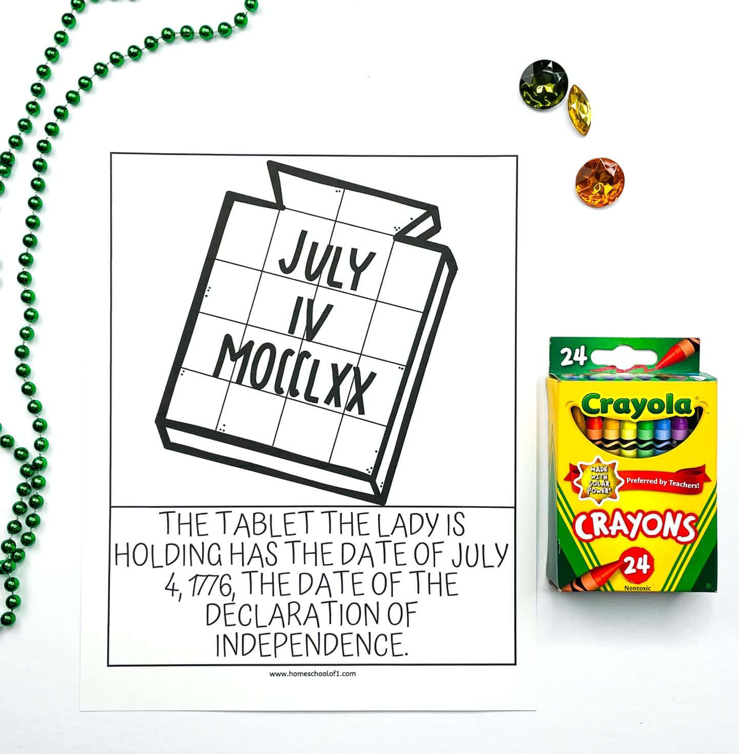 A black and white coloring worksheet showing the tablet held by the Statue of Liberty, inscribed with the date “JULY IV MDCCLXX.” Below the image, the caption reads: “The tablet the lady is holding has the date of July 4, 1776, the date of the Declaration of Independence.” The worksheet is set on a white background with a green beaded necklace, colorful plastic gems, and a box of Crayola crayons.